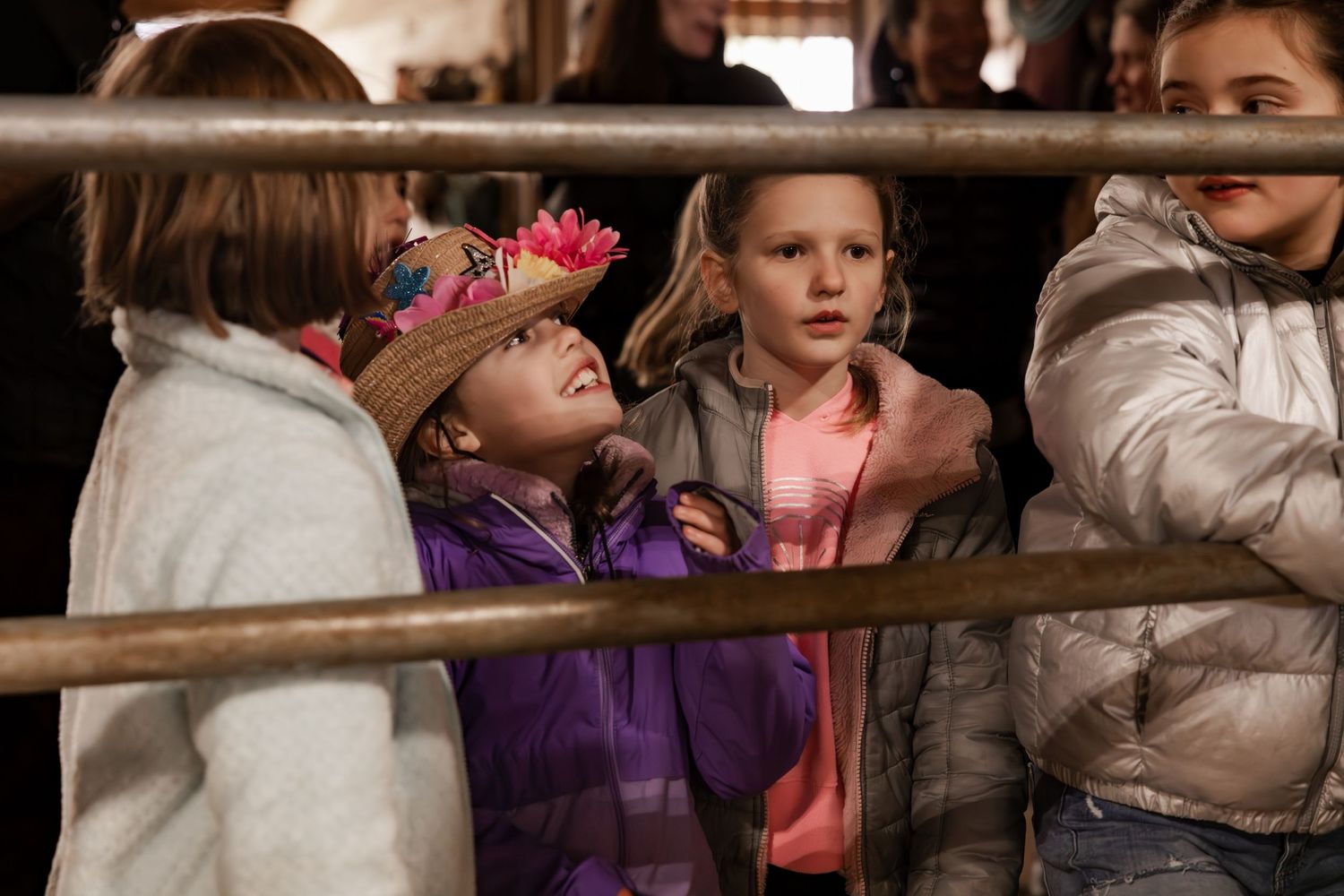 Children wrapped in winter coats peer through the wooden rails of a fence or barrier with curious expressions.