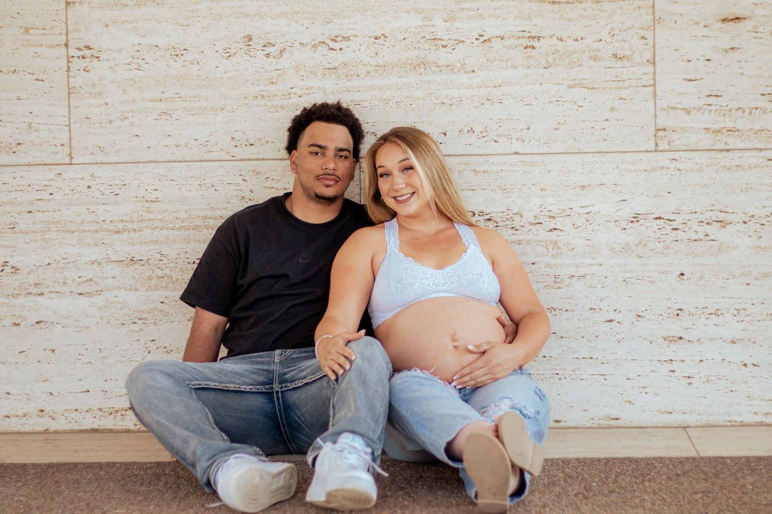 A couple sits together against a light colored wall, with the woman wearing a light blue top showing her pregnant belly.