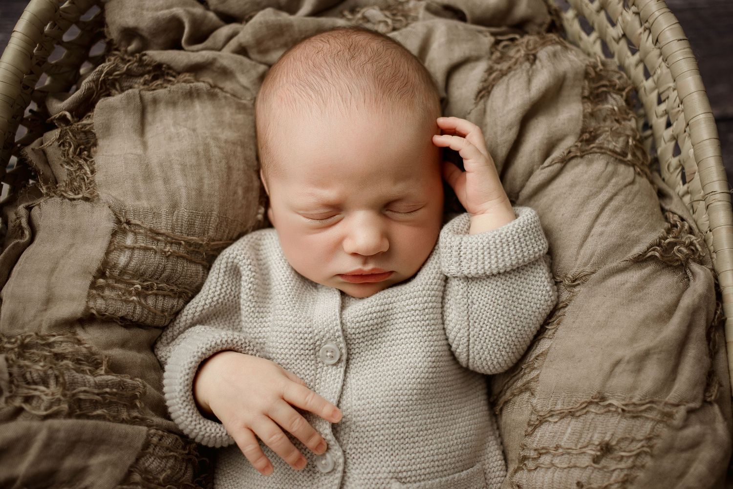 Sleeping newborn wearing cozy gray cardigan sweater rests peacefully on beige cushions.