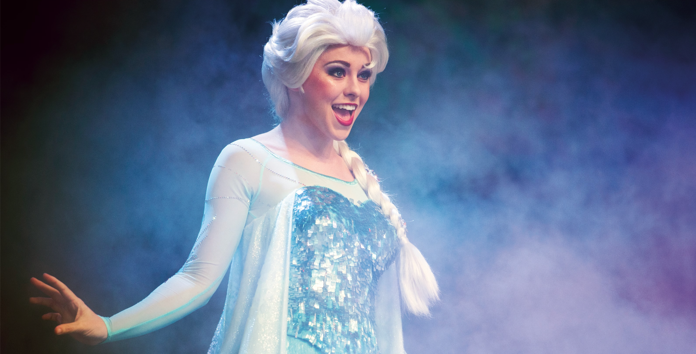 A performer in a glittering blue ice-queen costume poses with arms outstretched against a misty blue background.