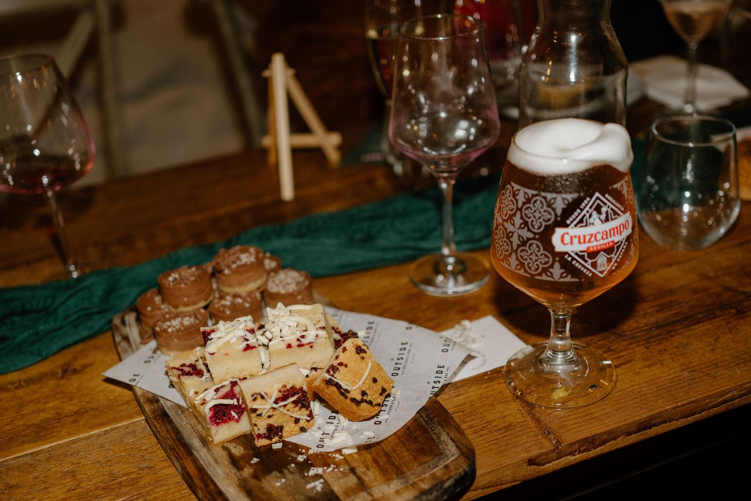A glass of dark craft beer sits on a wooden table next to appetizer plates and wine glasses.