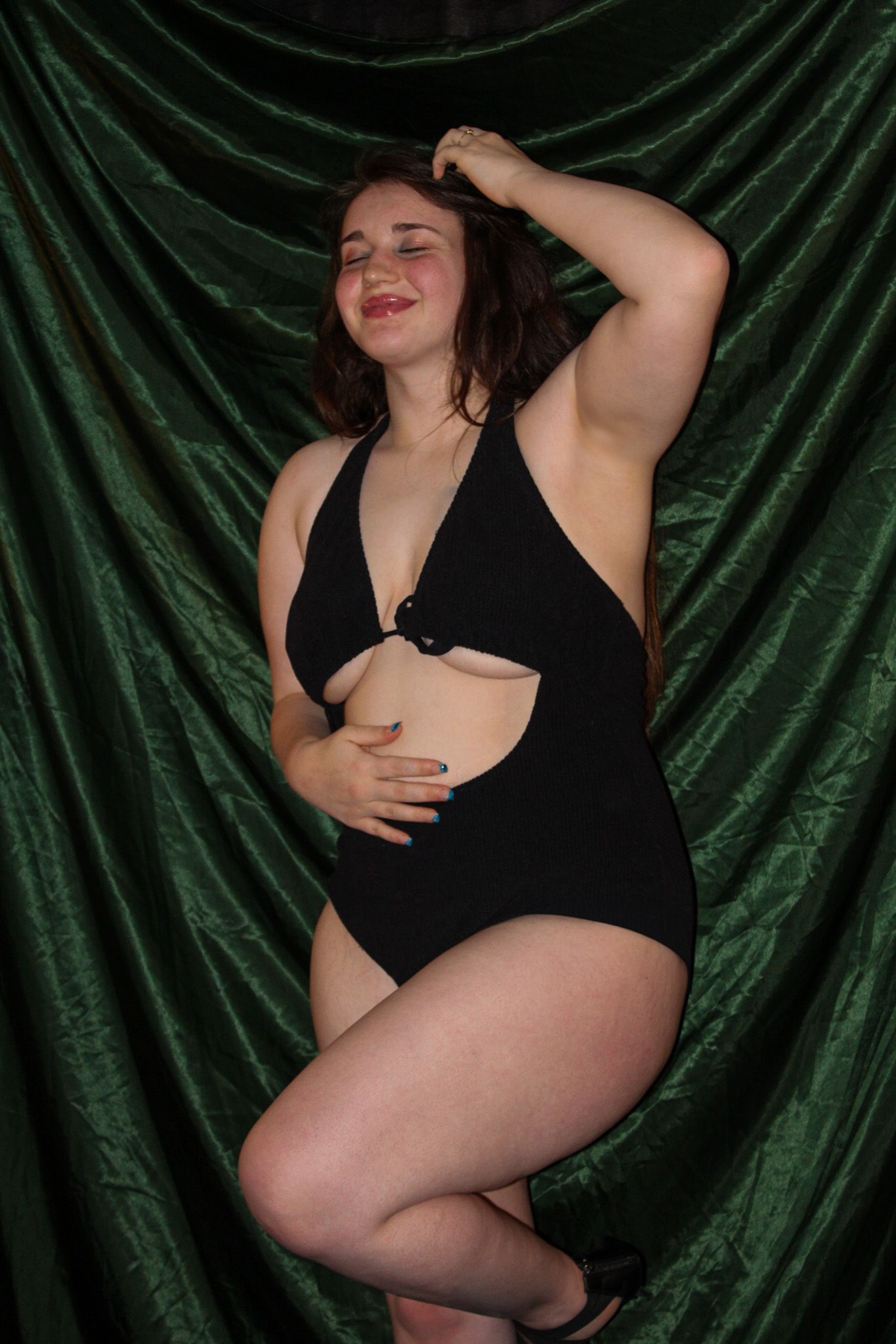 A person poses in a black swimsuit against a dark green velvet backdrop in a glamour photography setting.
