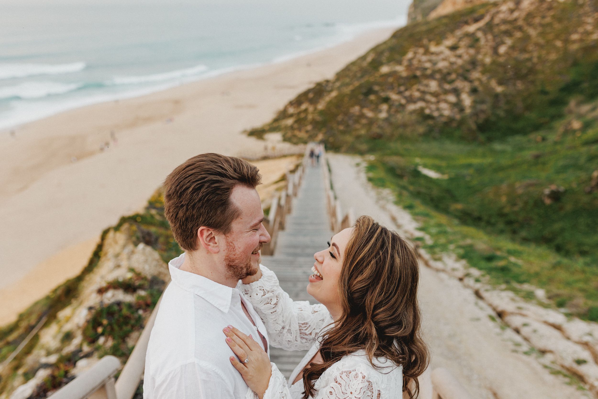 Two people embracing and laughing together on a scenic coastal path overlooking a sandy beach during a photoshoot in a beach near Lisbon