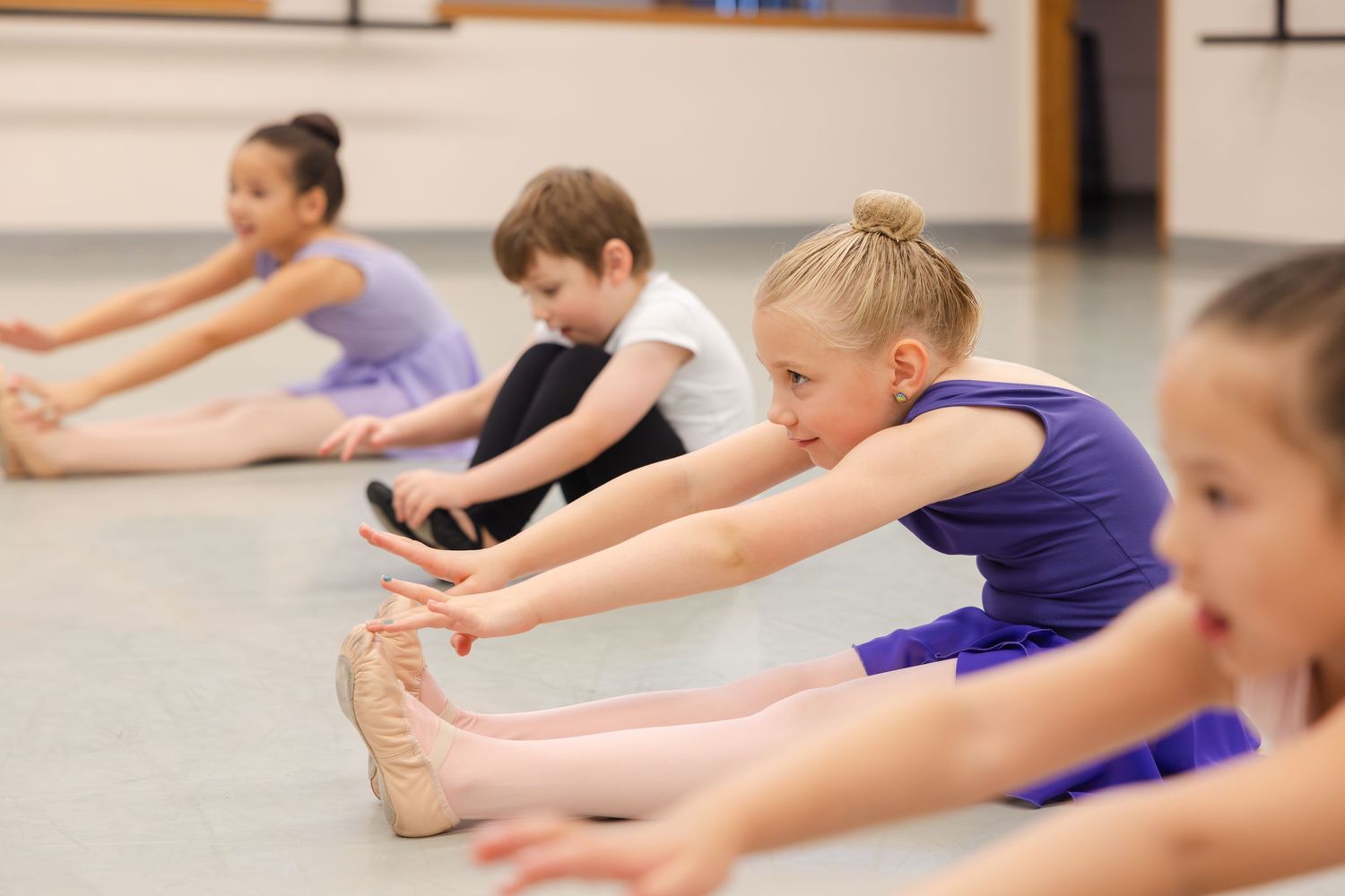 Young dancers in purple leotards stretch forward in a seated position during a ballet class.
