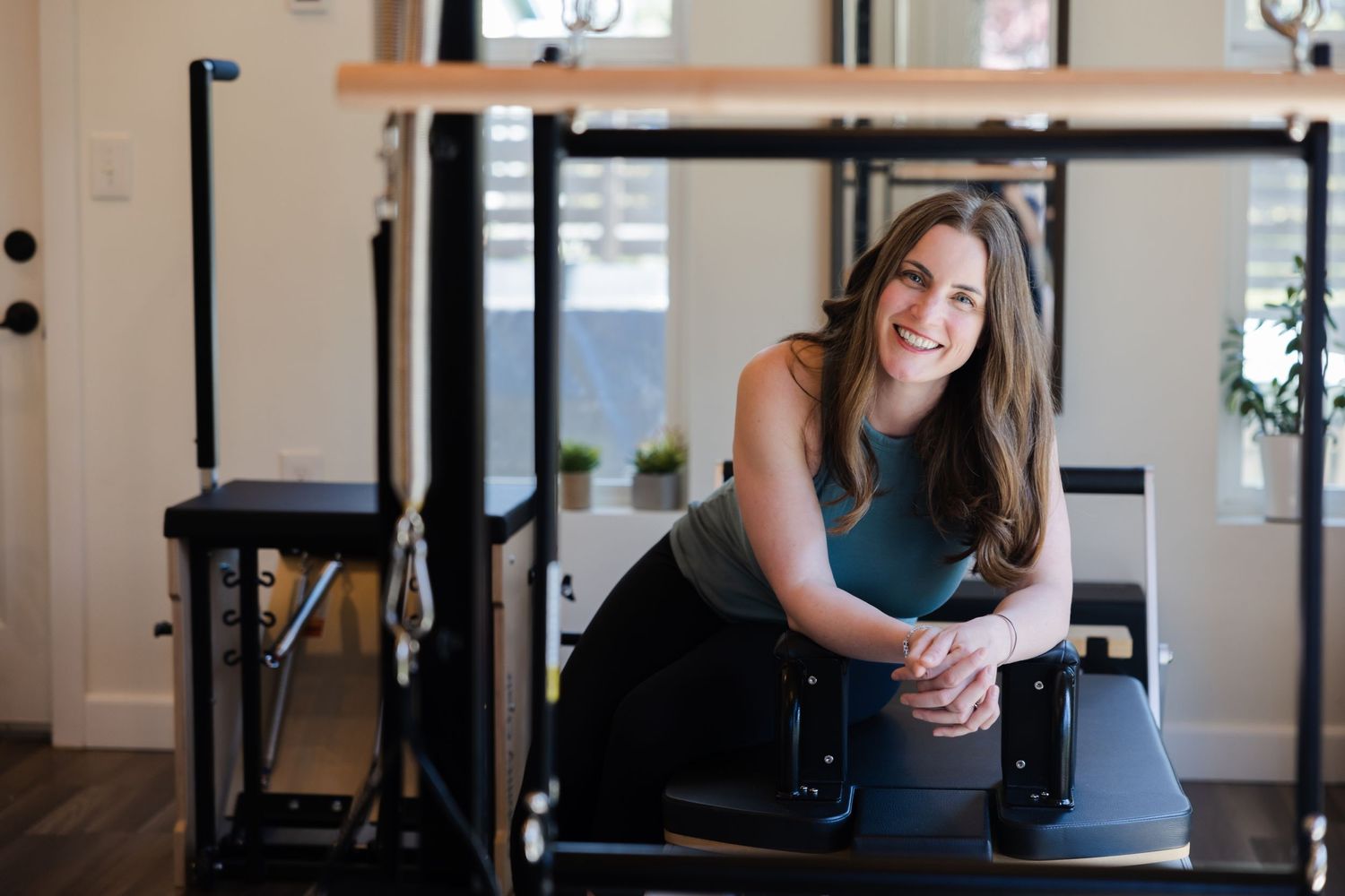 Fitness instructor sits on exercise equipment in a bright, modern pilates studio setting.