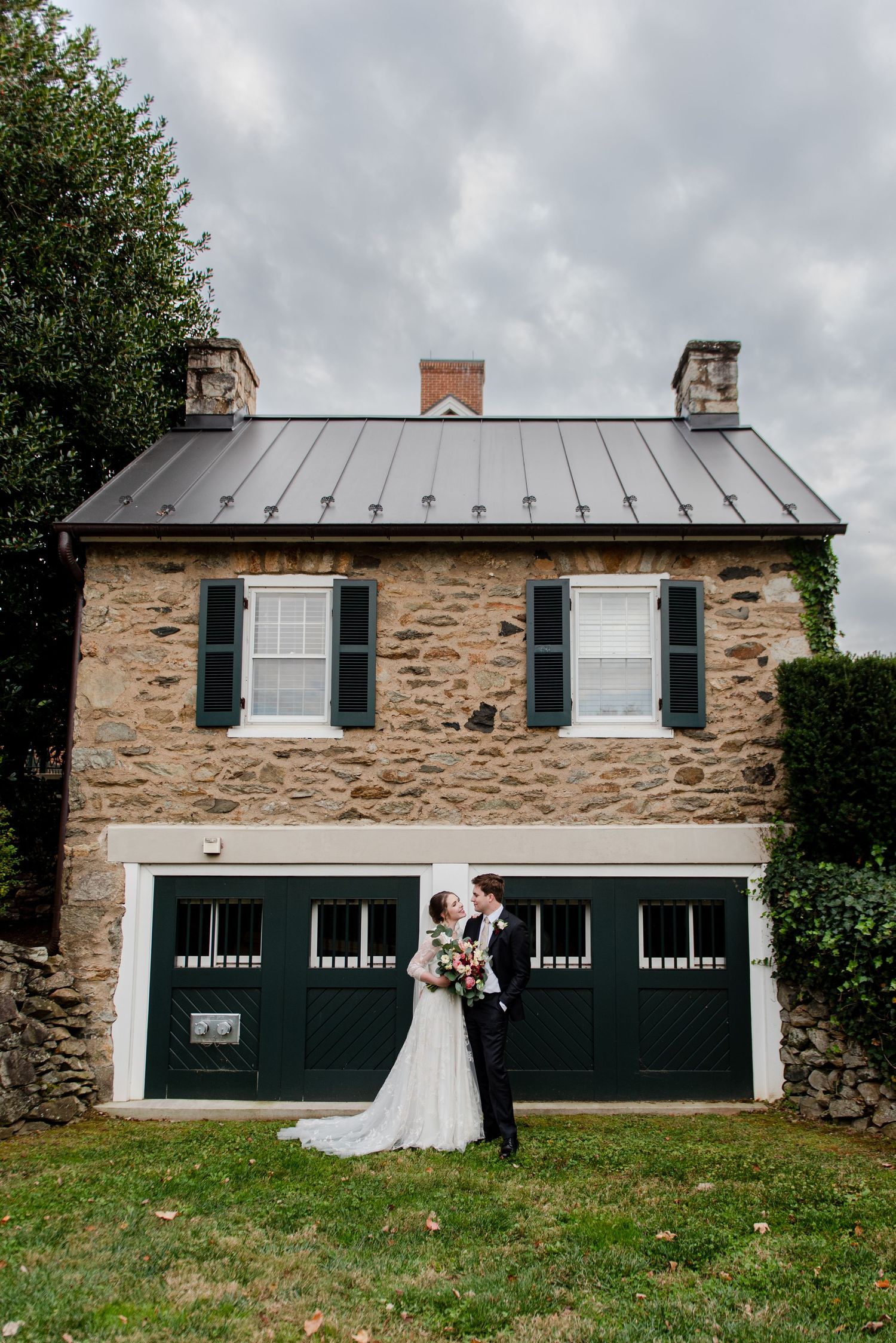 A rustic stone carriage house with black doors provides a charming backdrop for a wedding portrait. Middleburg Virginia