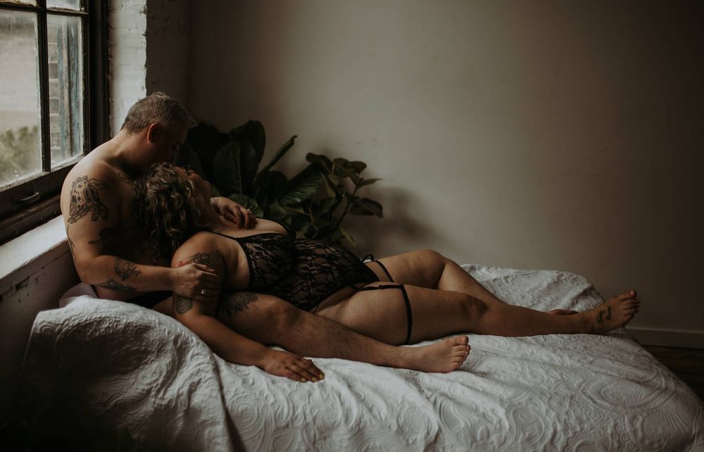 A couple shares an intimate moment together on a bed near a window with houseplants in moody lighting.
