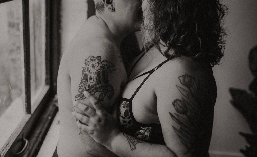 Black and white intimate couple embracing near window, showing tattoos and romantic connection.