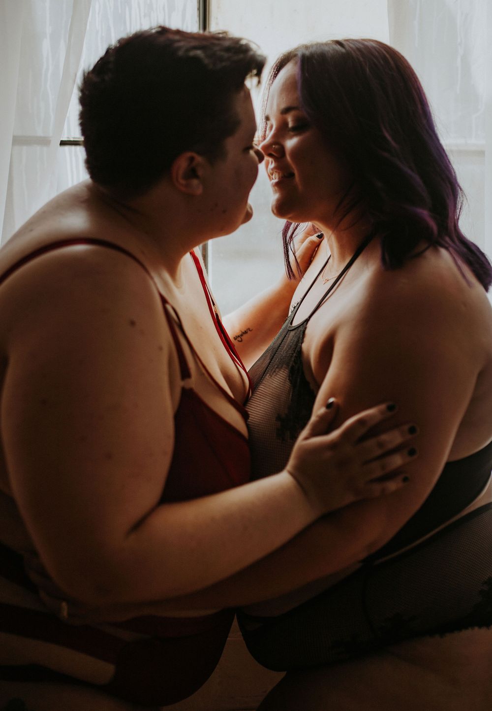 Two plus-size women in lingerie share an intimate romantic moment, embracing and leaning in for a kiss near a window.