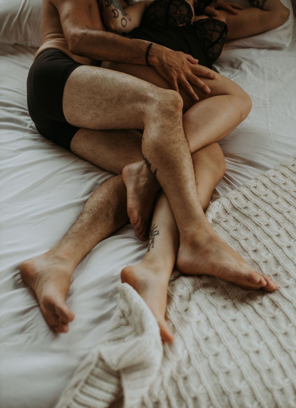 Intertwined bare legs of a couple lying intimately together on white bedding in a soft, romantic setting.