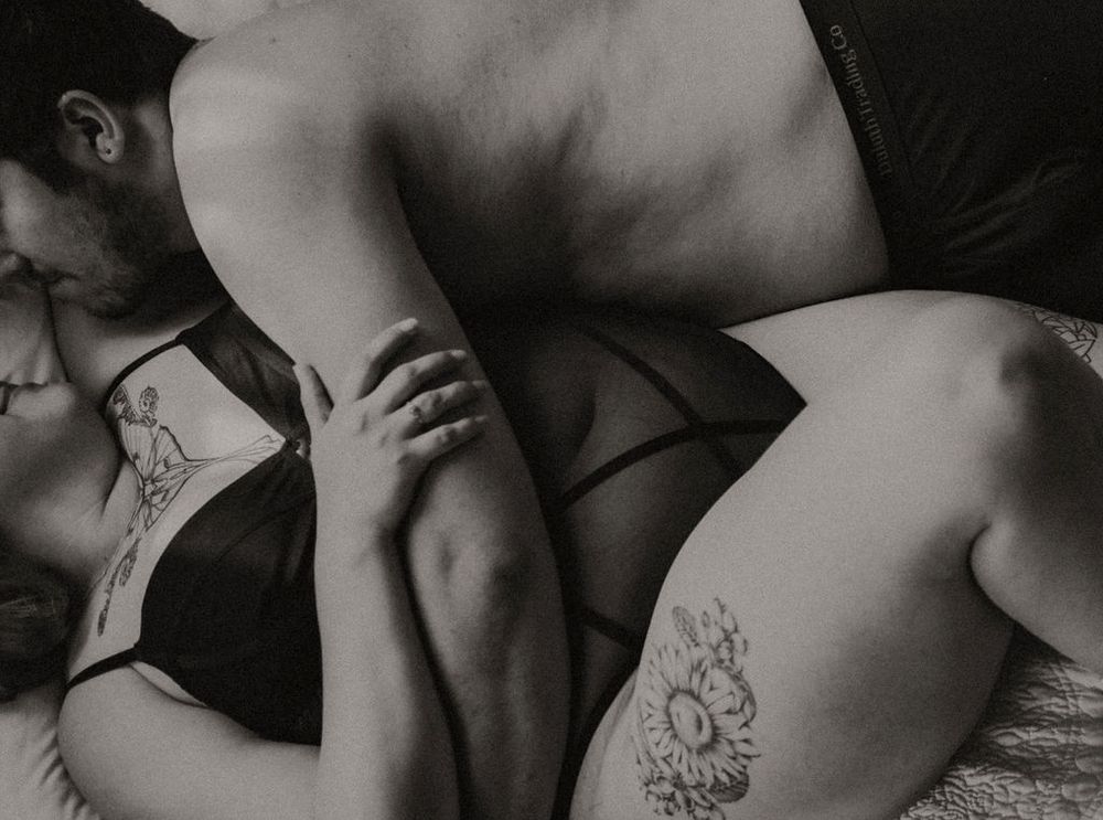Black and white intimate couple embracing, showing tattoos and lingerie in a romantic bedroom setting.