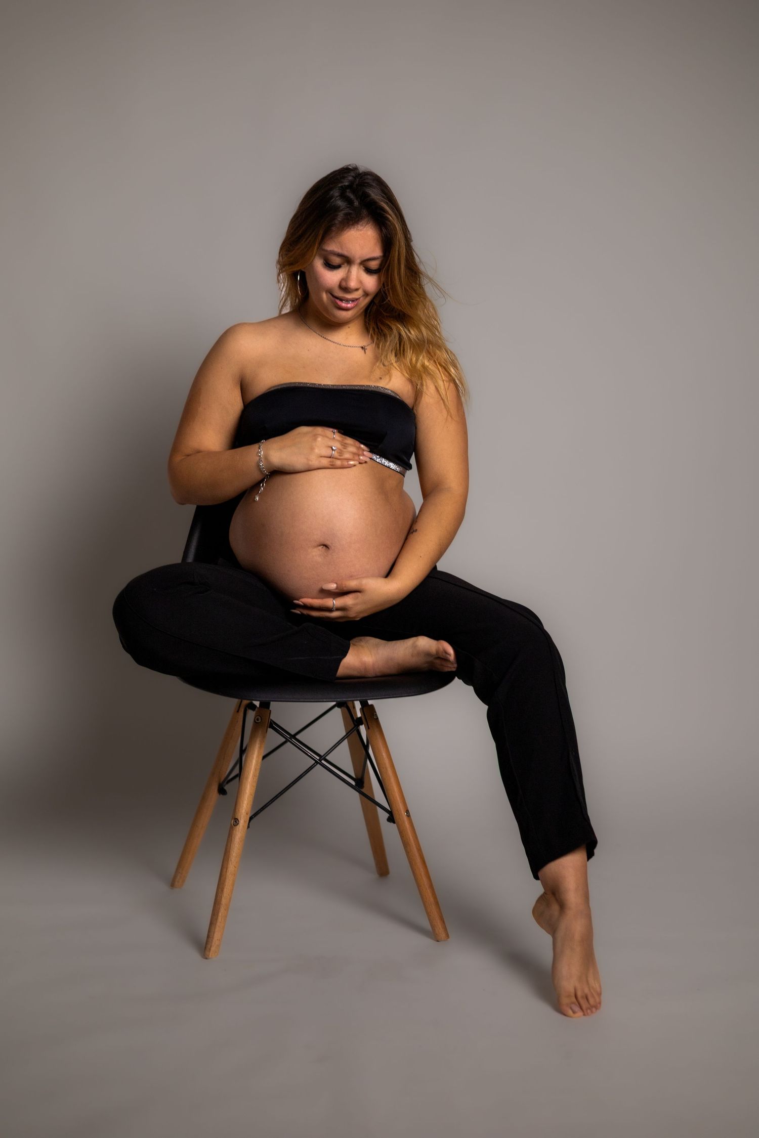 Minimalist maternity studio photos with woman in black outfit on wooden chair against grey backdrop.