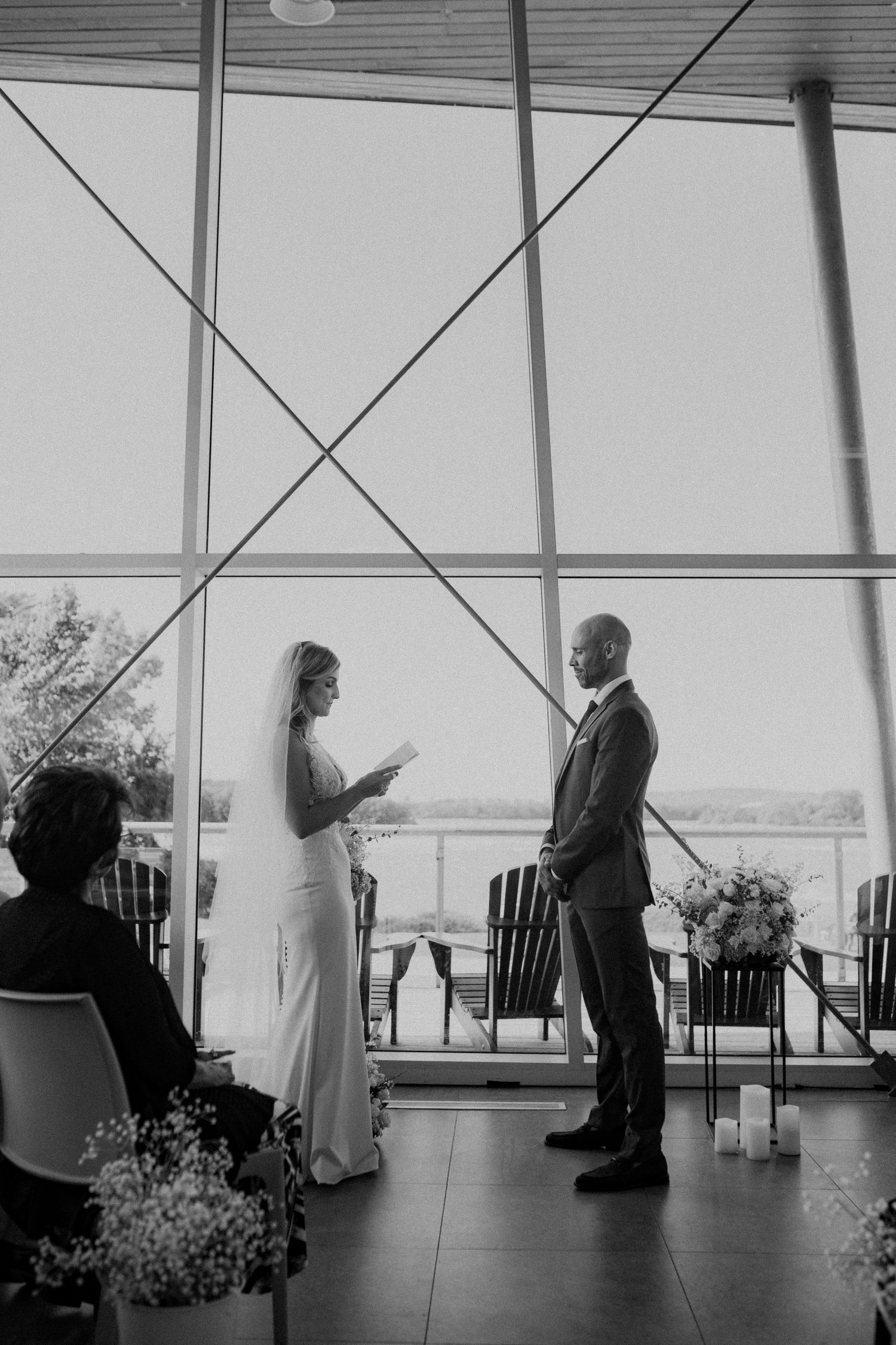 Black and white photo of bride reading vows to groom before large glass windows at elegant wedding ceremony.