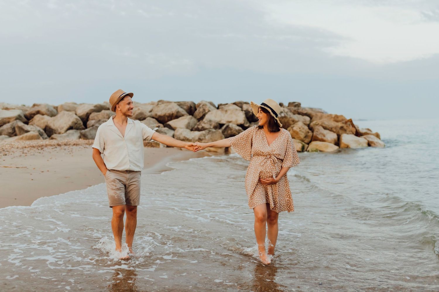 A maternity couple walking hand in hand through shallow water on Bournemouth beach.