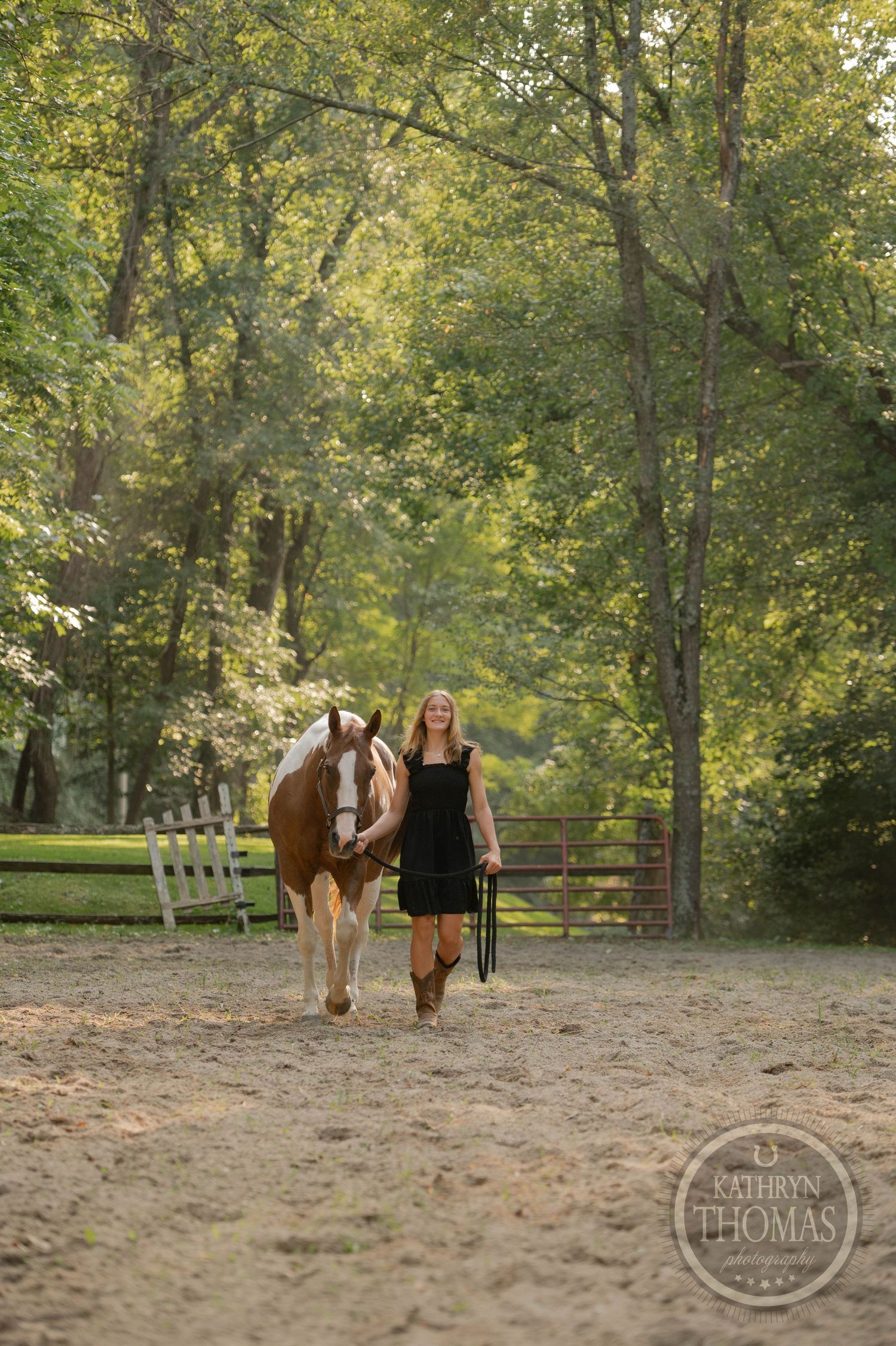 A person walks alongside their paint horse on a dirt path through trees and fencing.
