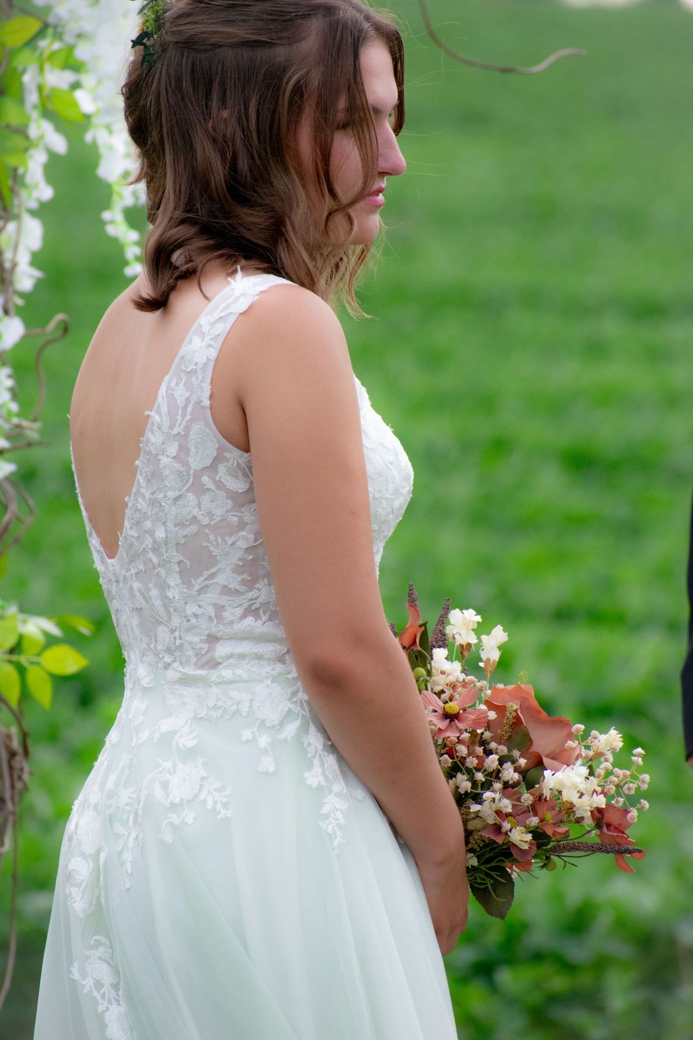 Back view of a bride in a white lace wedding dress holding a pink and white floral bouquet in a garden setting.