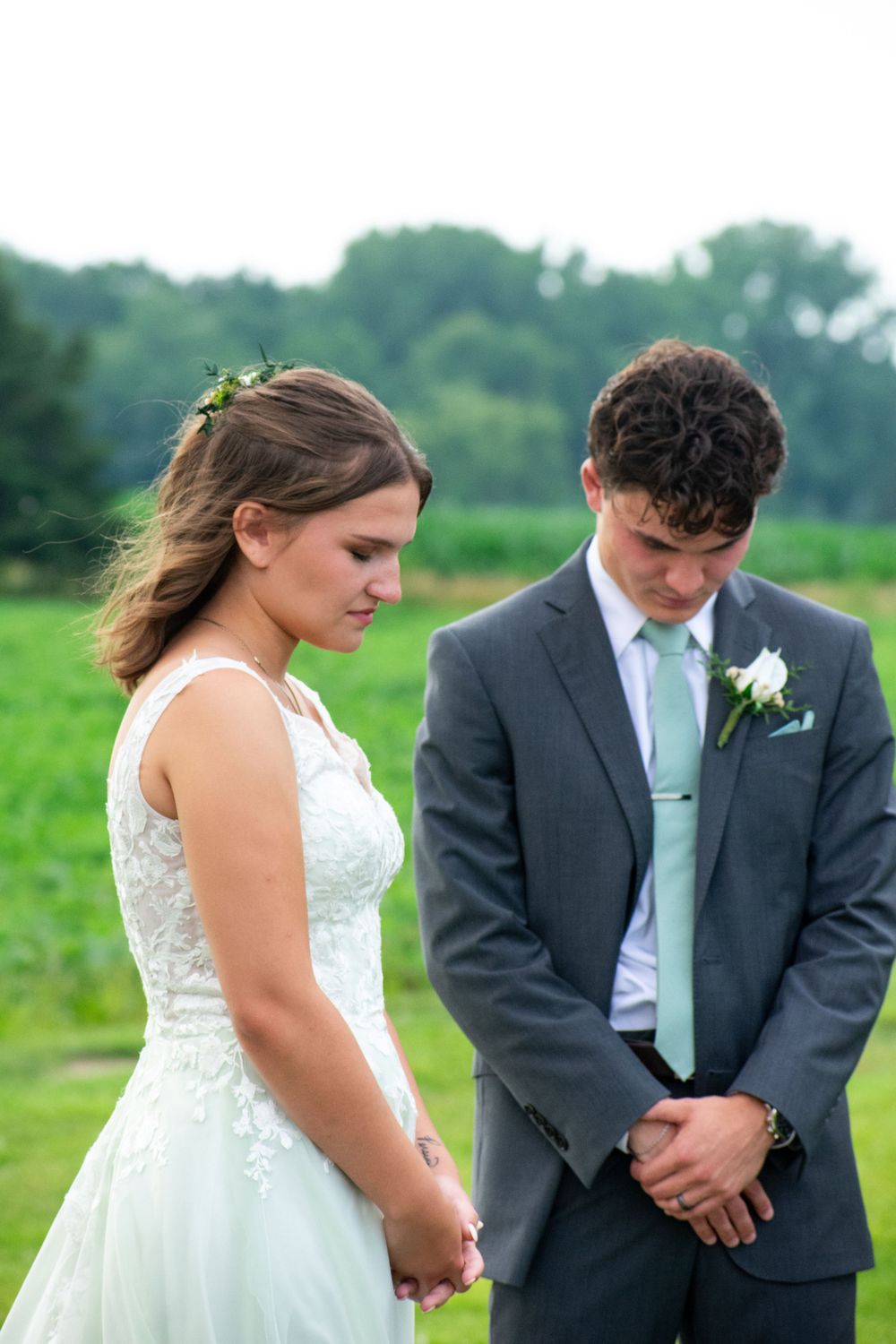 A solemn wedding ceremony moment takes place outdoors against a backdrop of green trees.