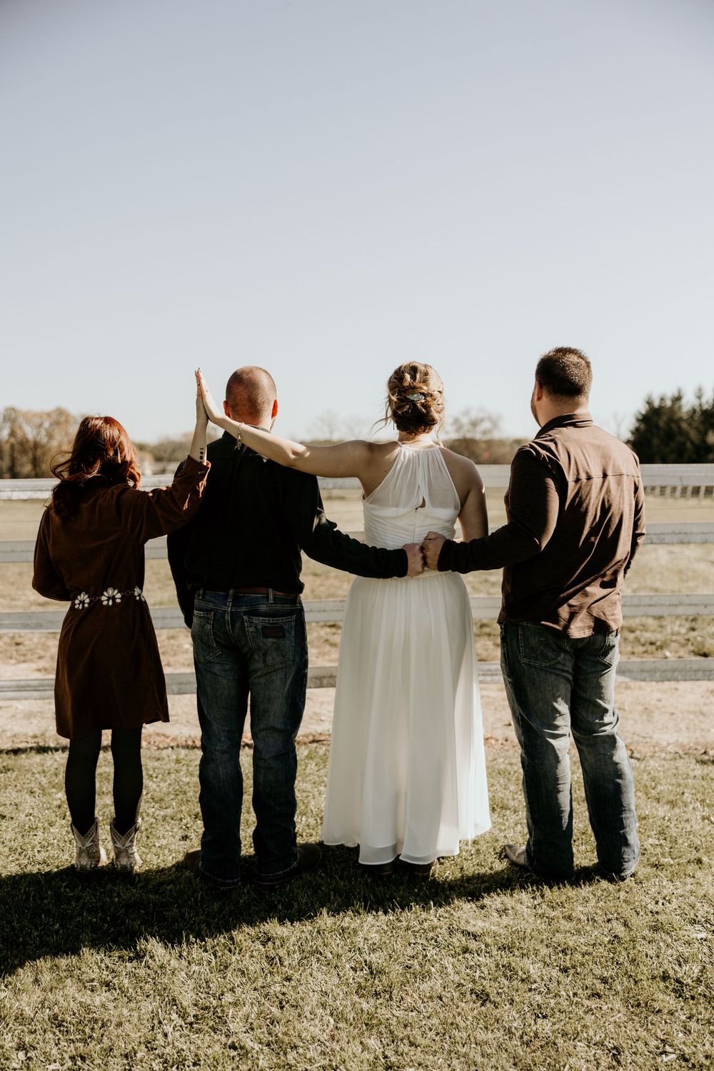 Back view of four people standing together on grass with one person wearing a white dress in the middle.