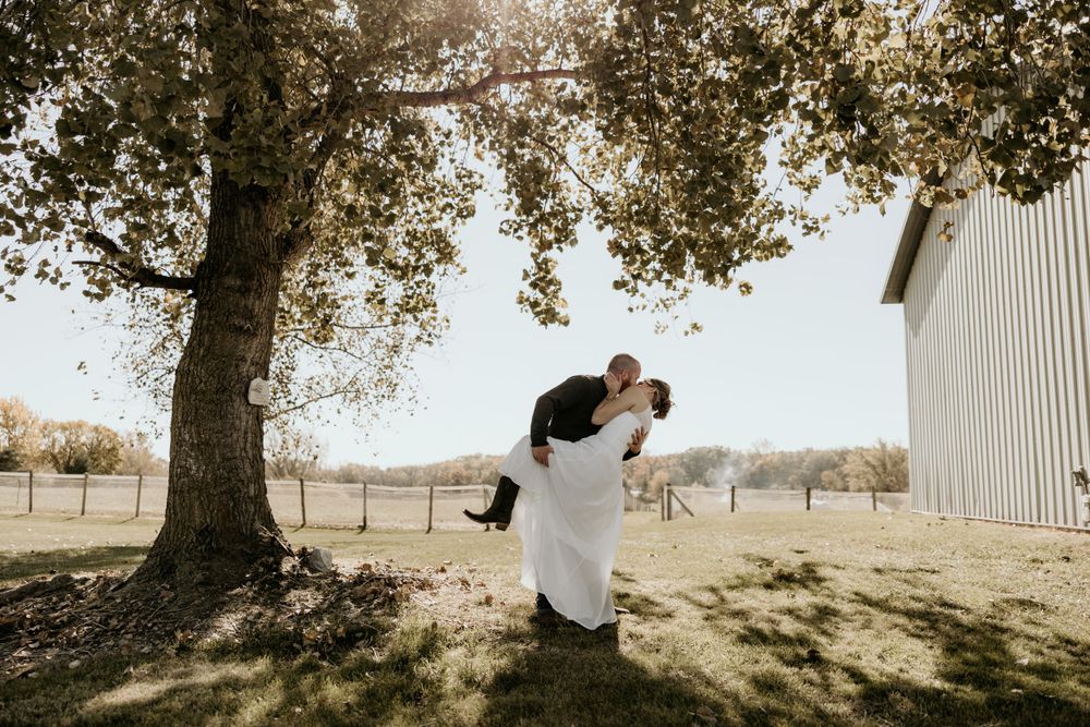 A couple embraces under a large tree beside a rustic barn on their wedding day.