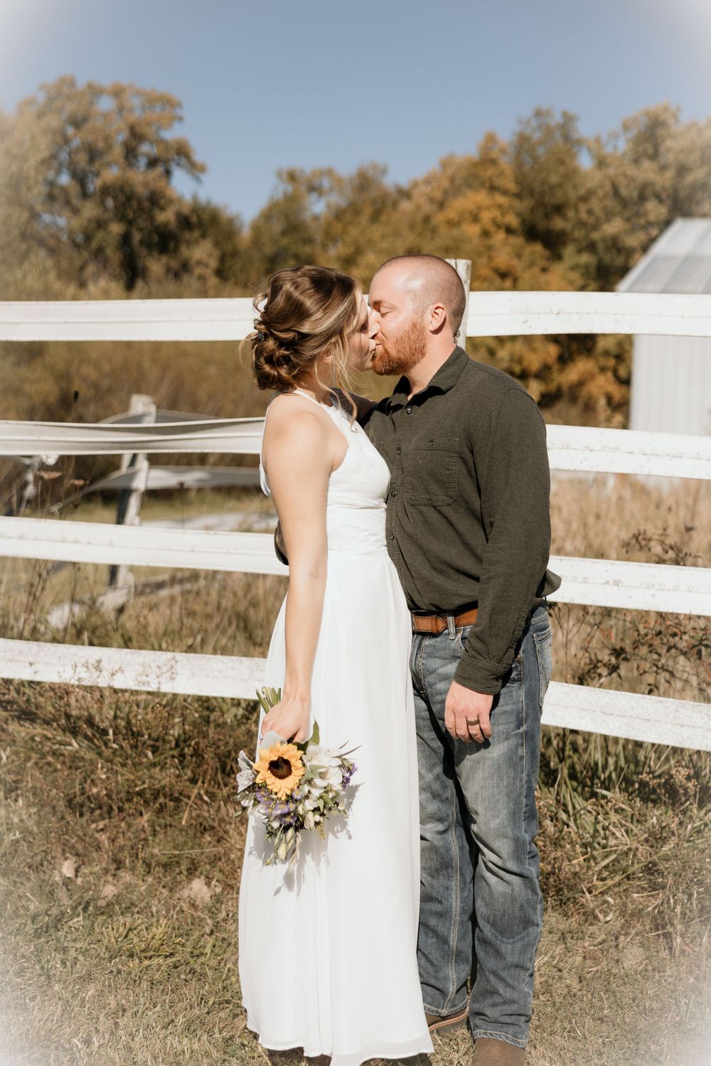 A romantic couple shares a kiss by a white fence on their wedding day, with sunflowers in the bride's bouquet.