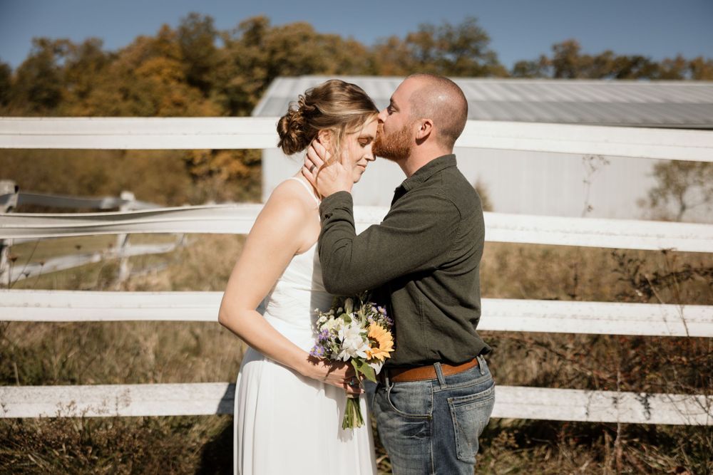 A couple embraces in front of a white fence on a sunny autumn day with fall foliage in the background.