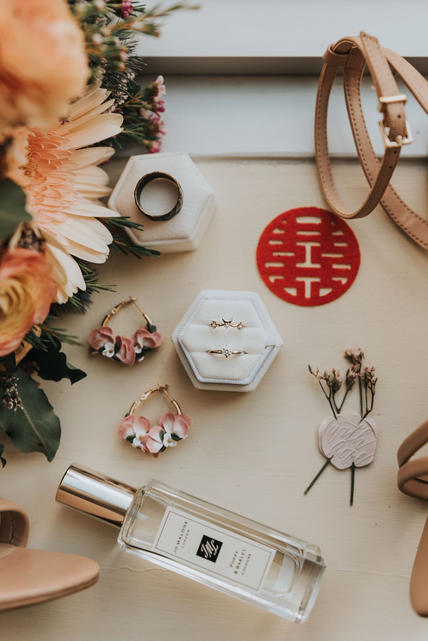 Wedding details flatlay featuring flowers, rings, perfume bottle, and delicate accessories on a neutral surface.