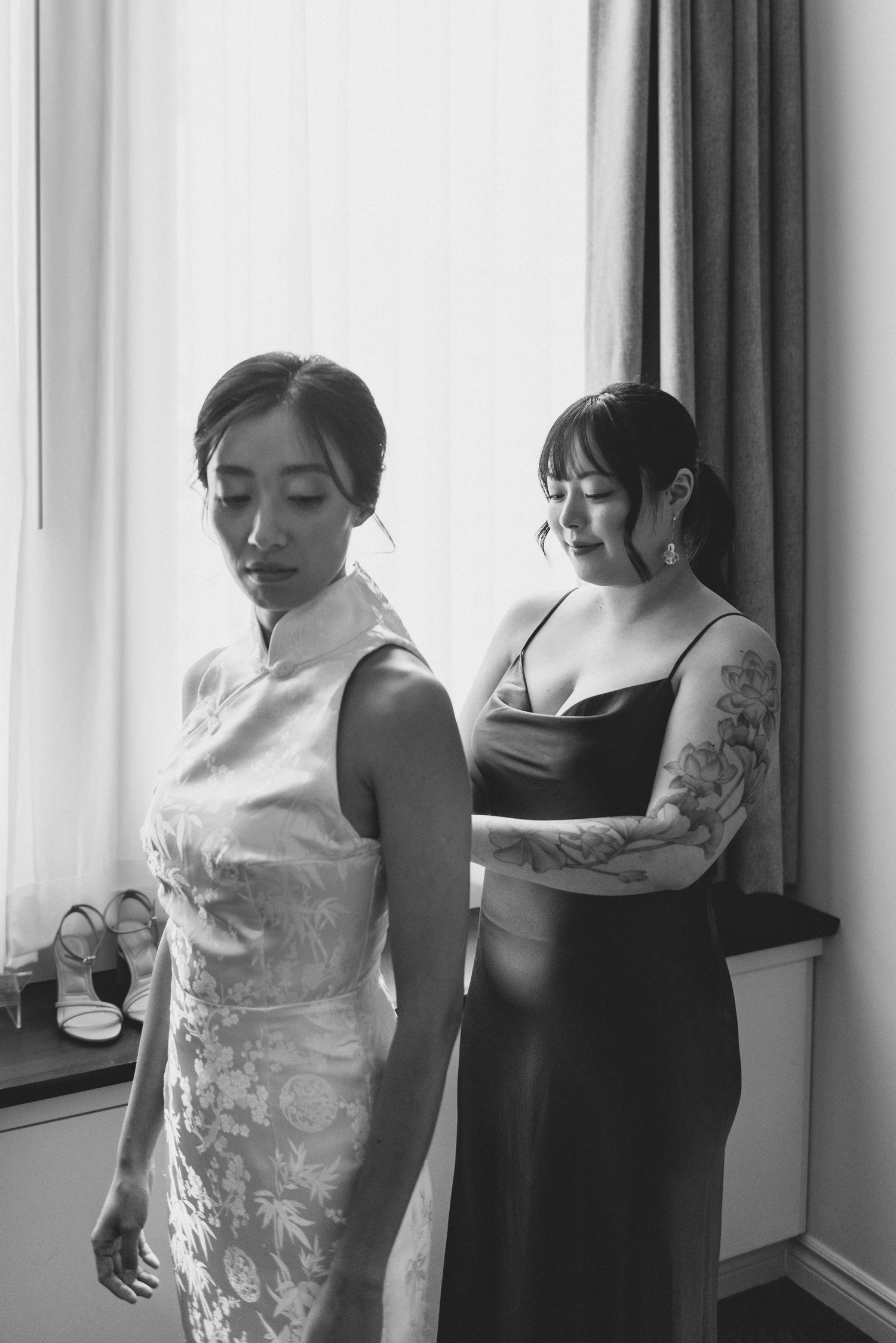 Black and white photo of two women in formal dresses standing by a window, one in a cheongsam and one in a dark gown.