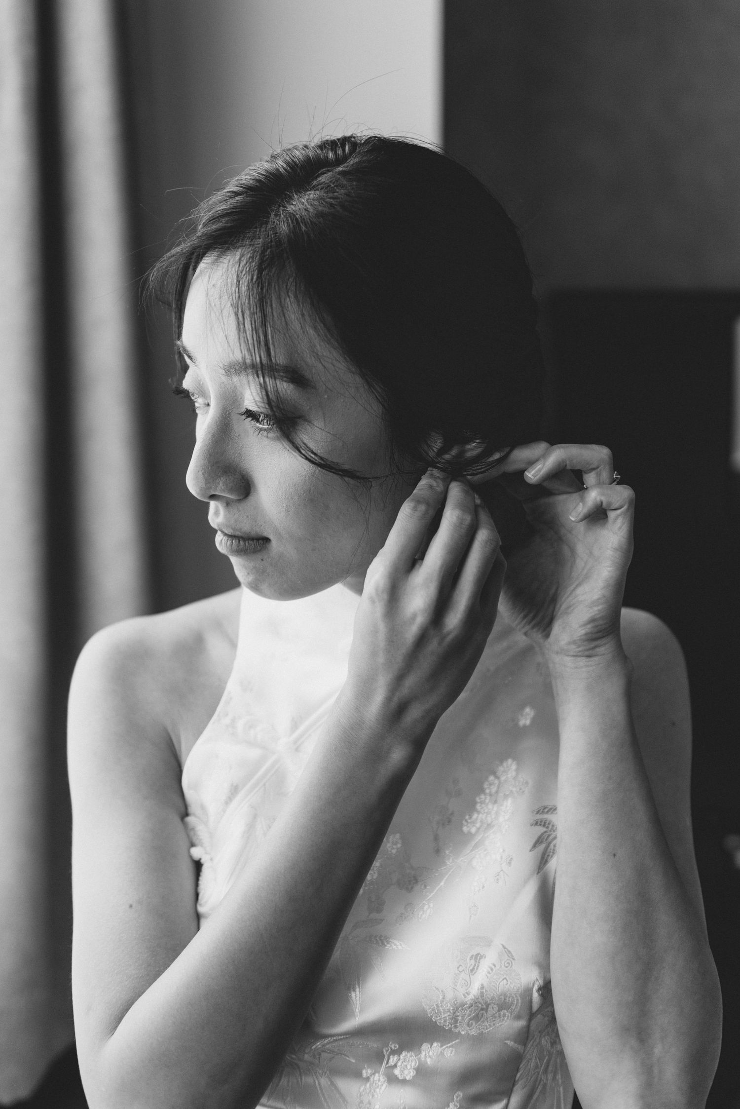 Black and white portrait of a bride in a white dress adjusting her hair near a window, captured in profile.