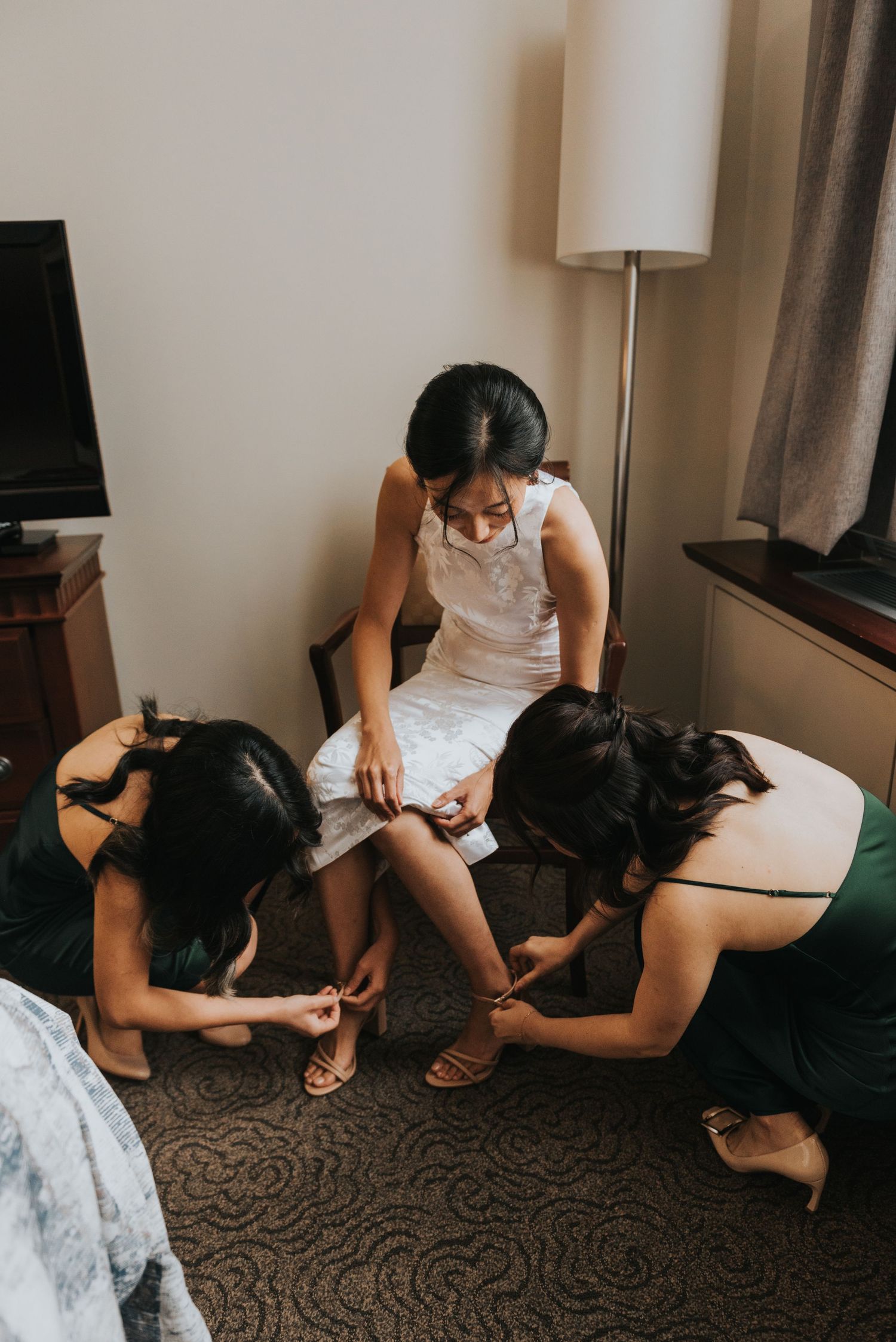 Wedding preparations show women helping with shoes on a carpeted floor with a floor lamp and TV stand visible.