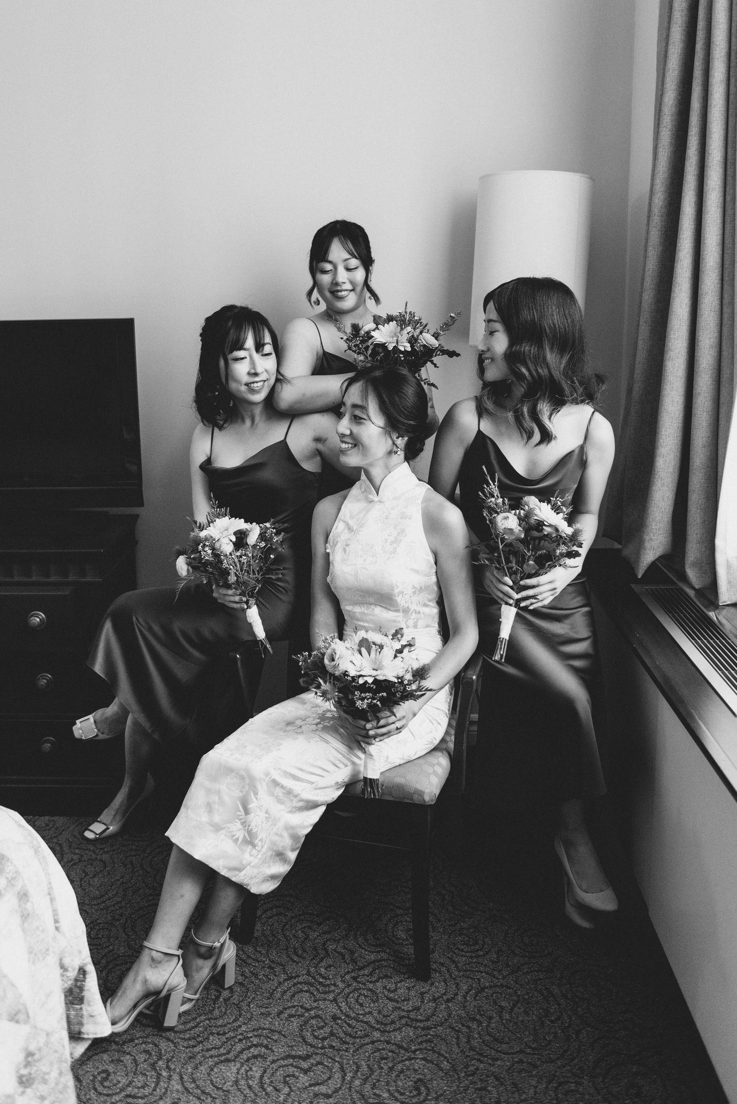A black and white photo of a bridal party in formal dresses sharing a candid moment together while holding bouquets.