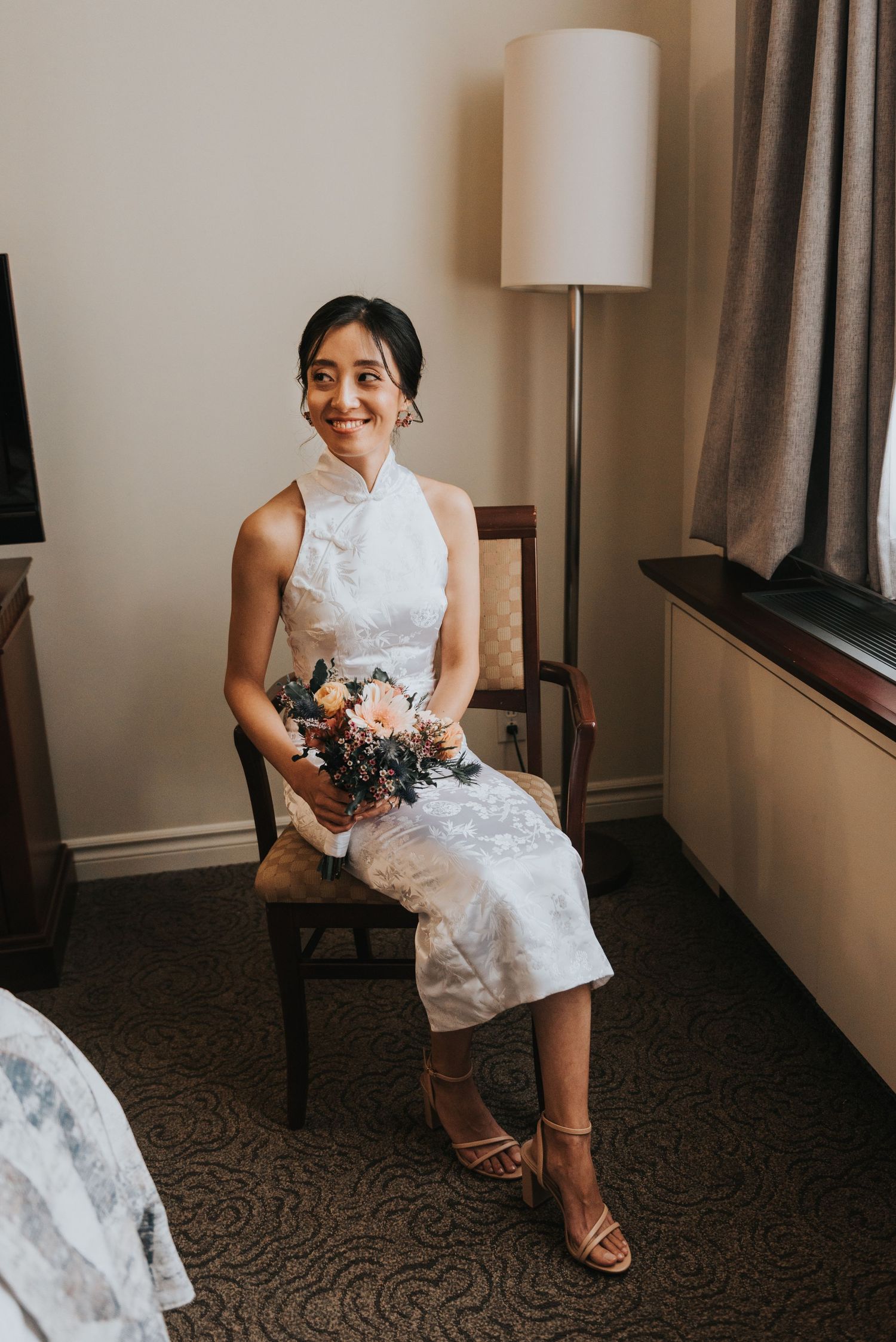 A bride in a white halter dress sits on a wooden chair holding a bouquet in an elegant indoor setting.