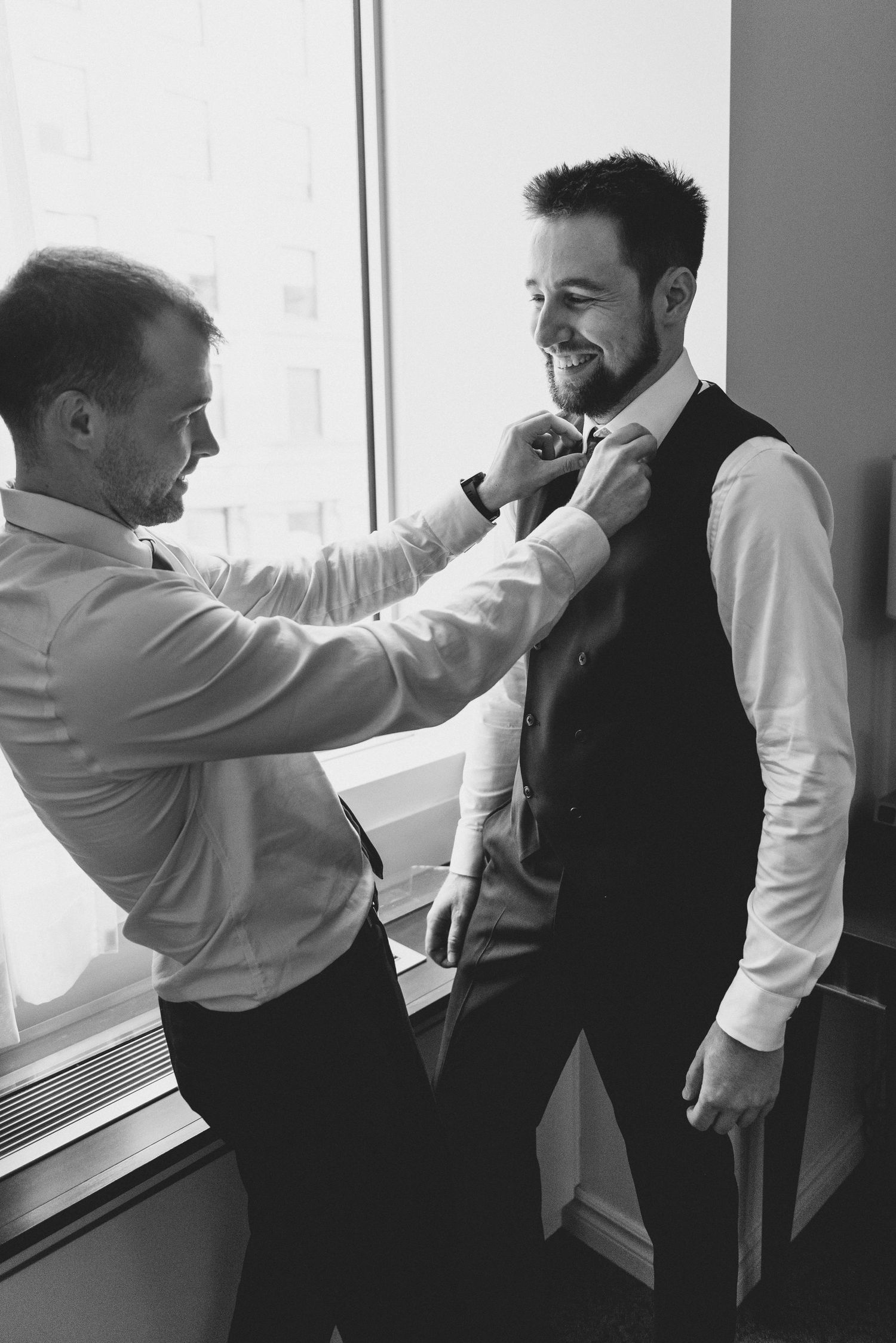 Two men share a candid moment while adjusting formal attire near a window in black and white photography.