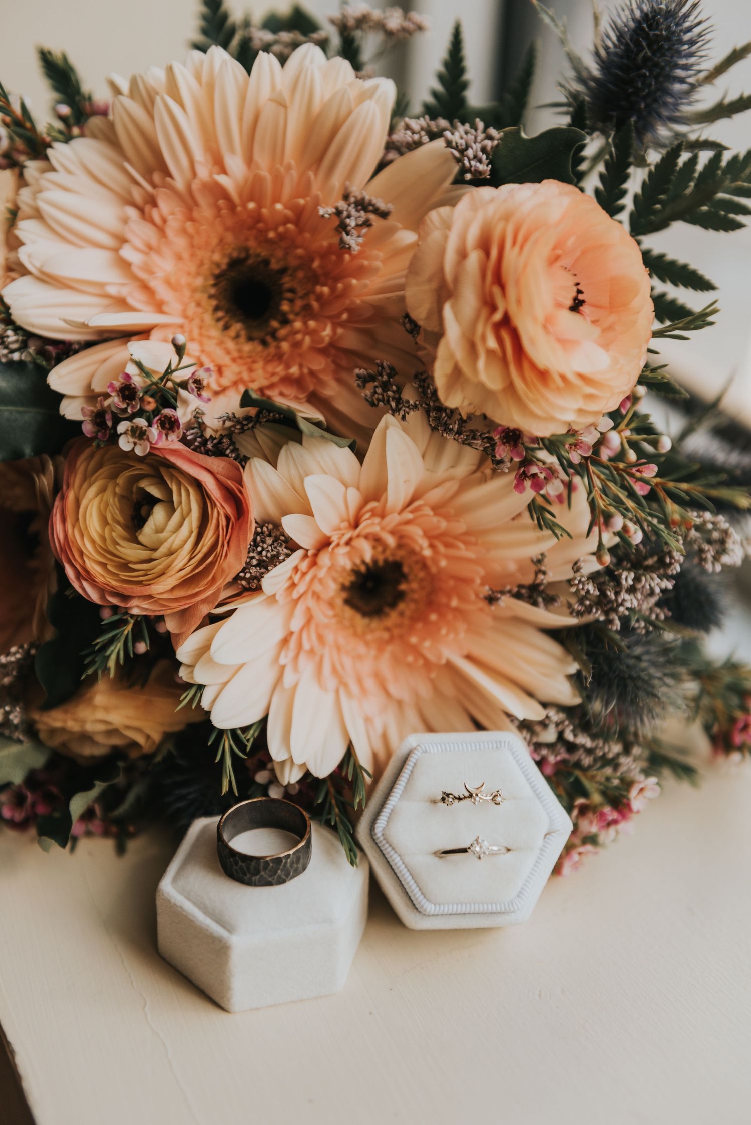 A peachy wedding bouquet with gerbera daisies and ranunculus flowers next to hexagonal ring boxes on a light surface.