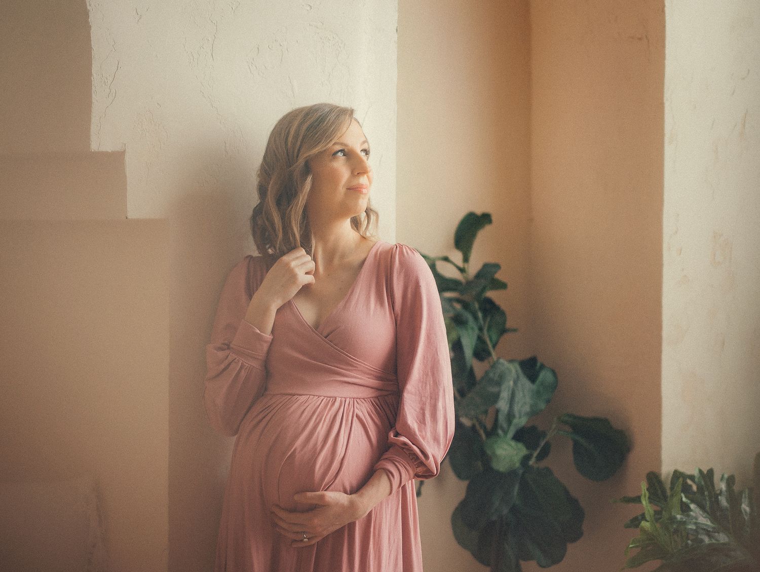 A person in a flowing pink maternity dress stands near a window with indoor plants, bathed in soft natural light.