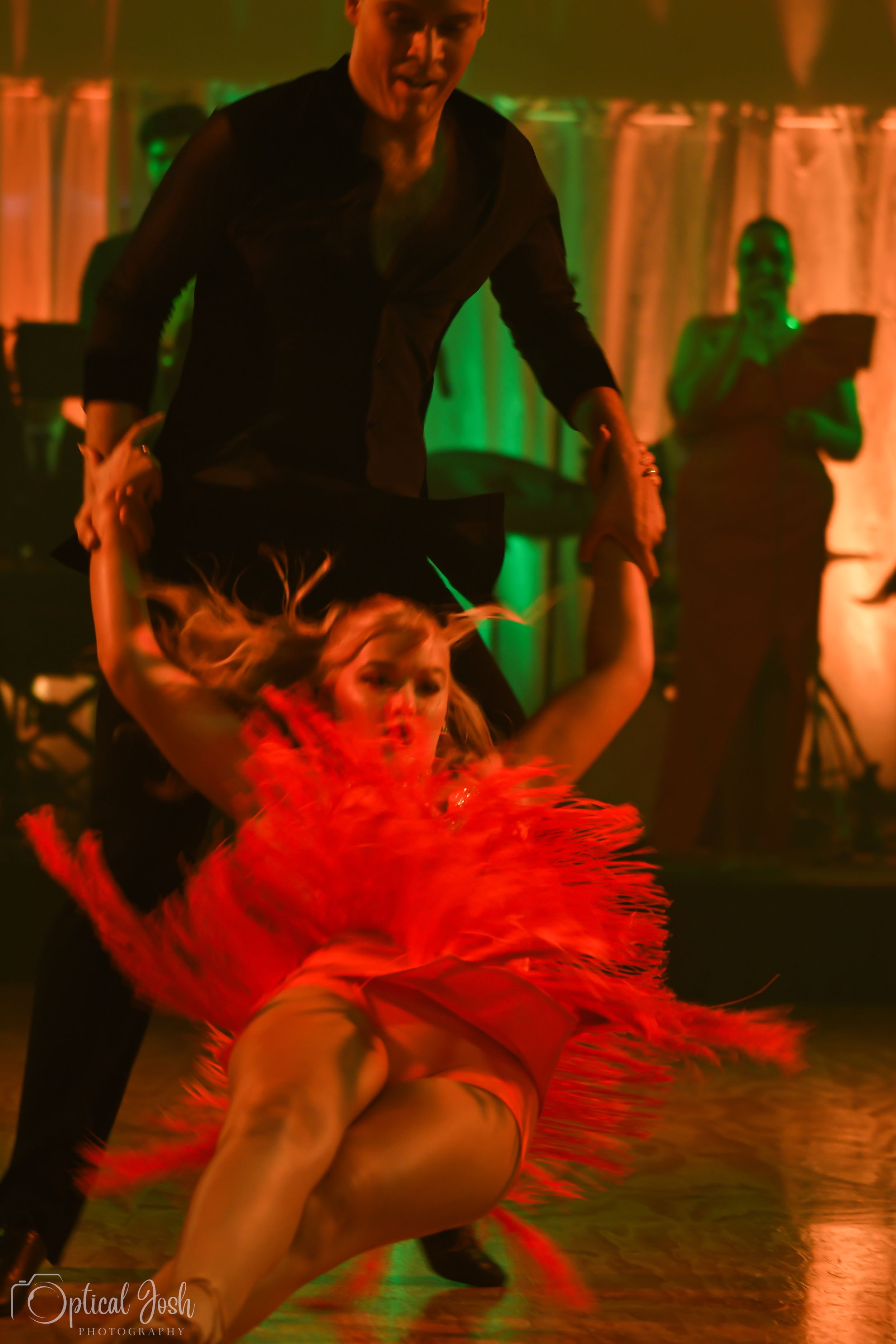 Ballroom dancers perform a dramatic dip move with flowing red costume under colorful stage lighting.