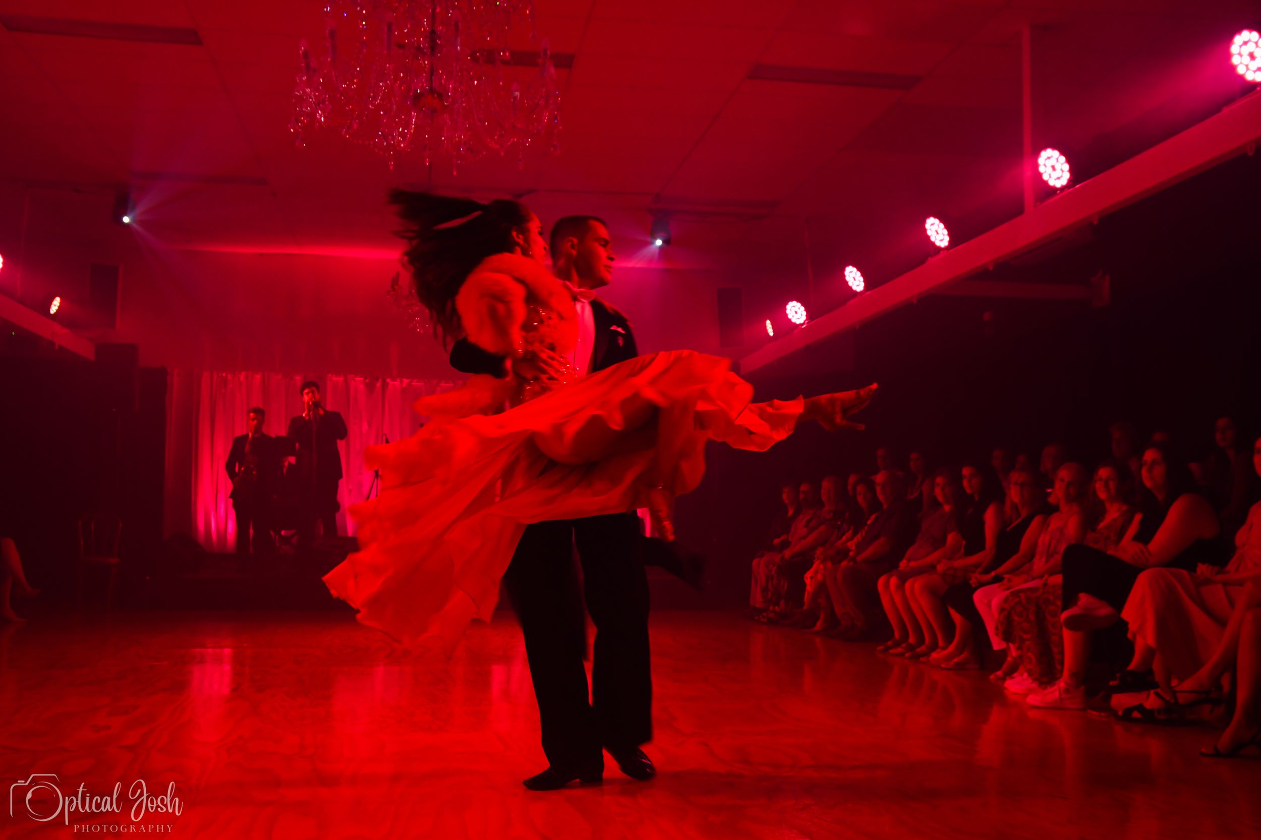 Dancers perform an elegant lift under dramatic red stage lighting as audience members watch from their seats.
