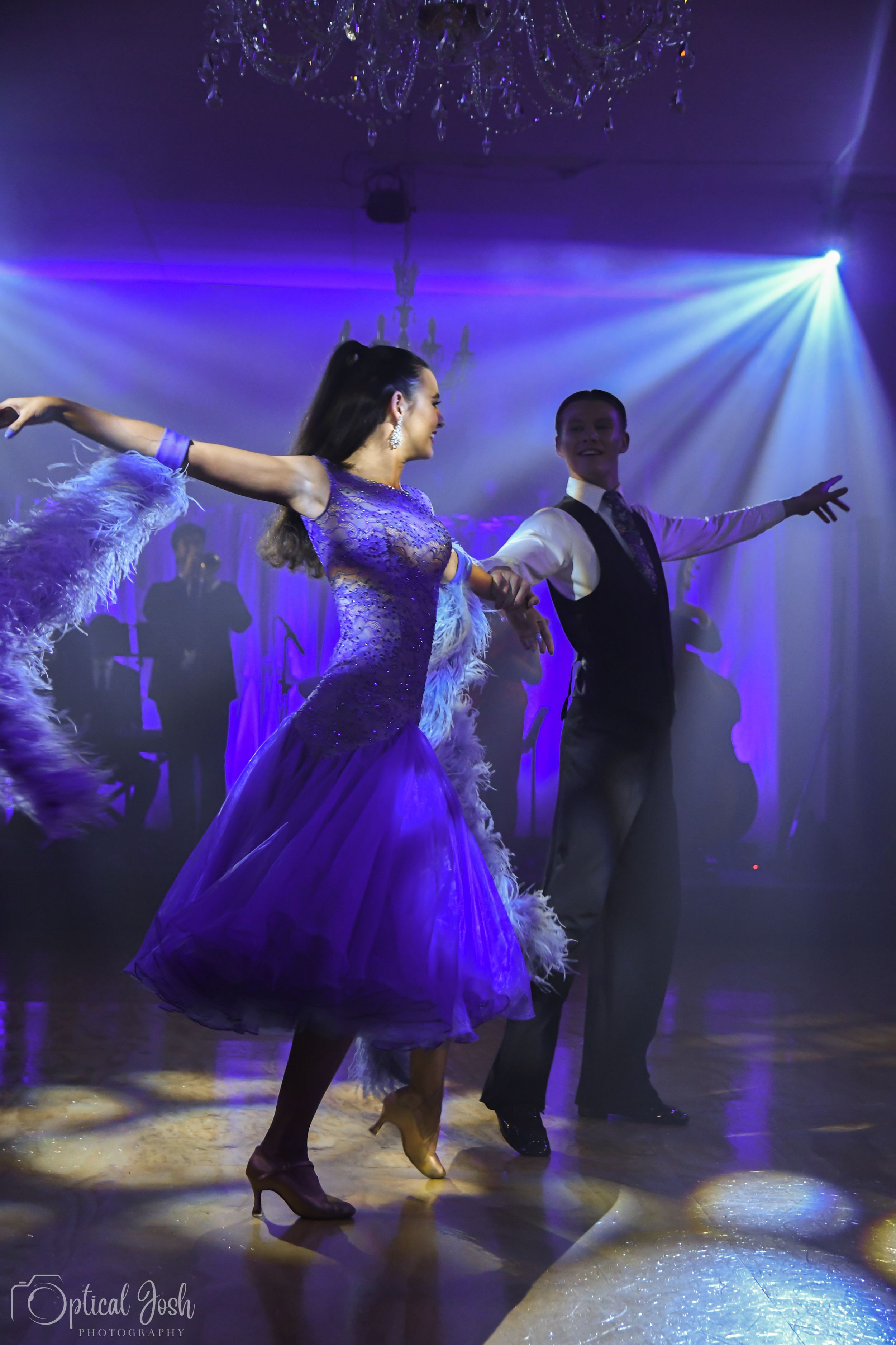 A couple performs an elegant ballroom dance in flowing purple gowns under dramatic blue stage lighting.