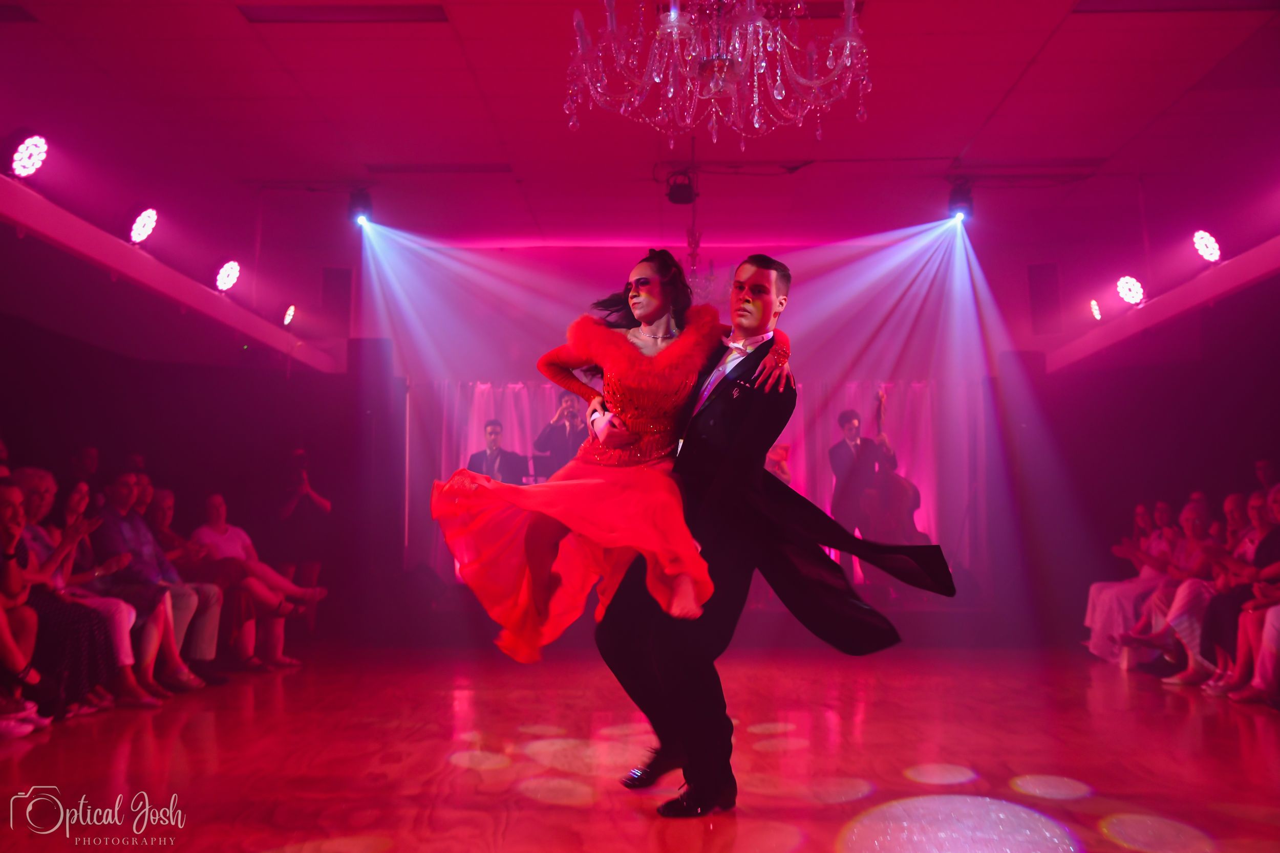 Two dancers perform a passionate tango under dramatic red lighting on a dance floor with white drapes and audience members.