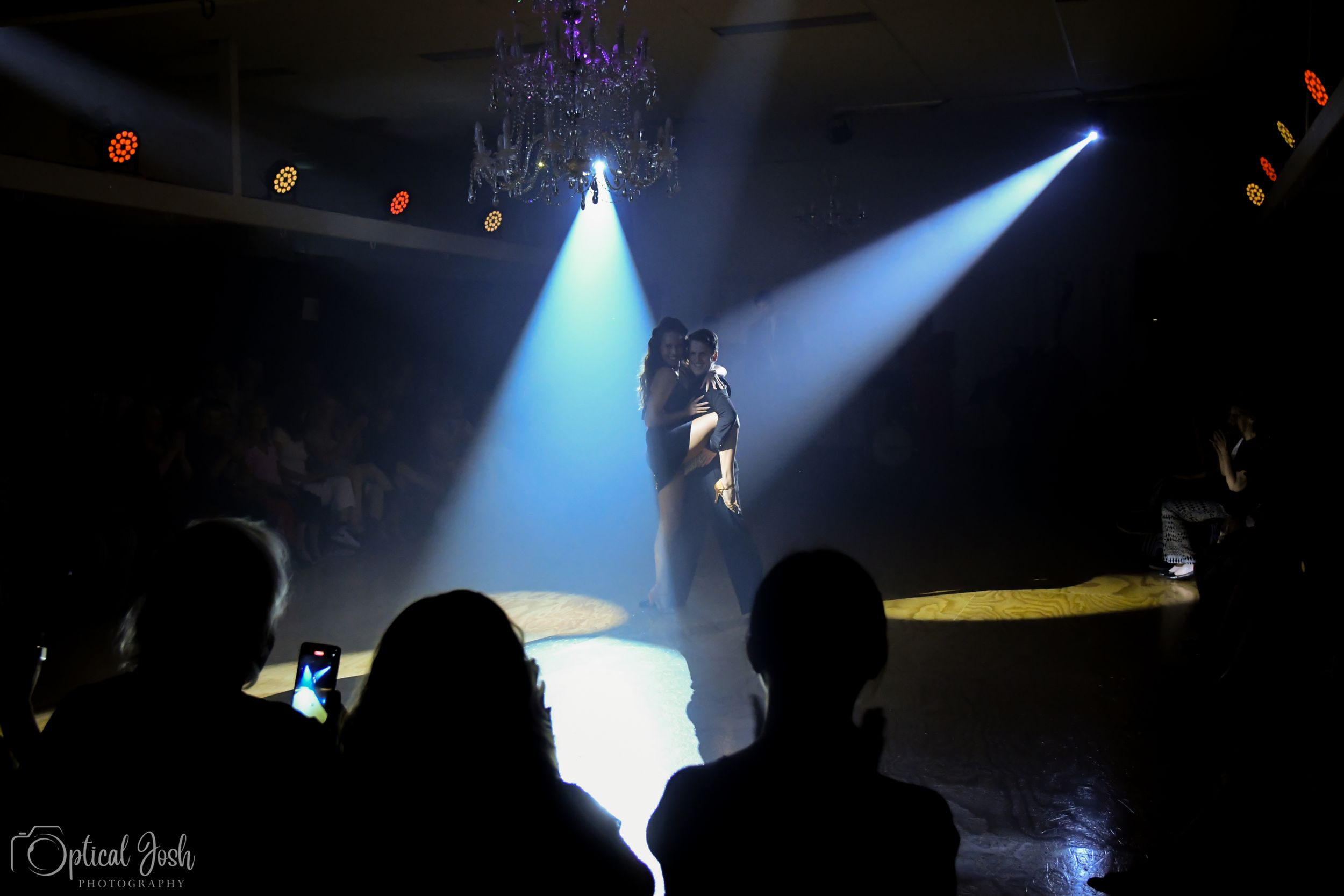 Performer on stage backlit by blue spotlights with audience silhouettes in foreground.
