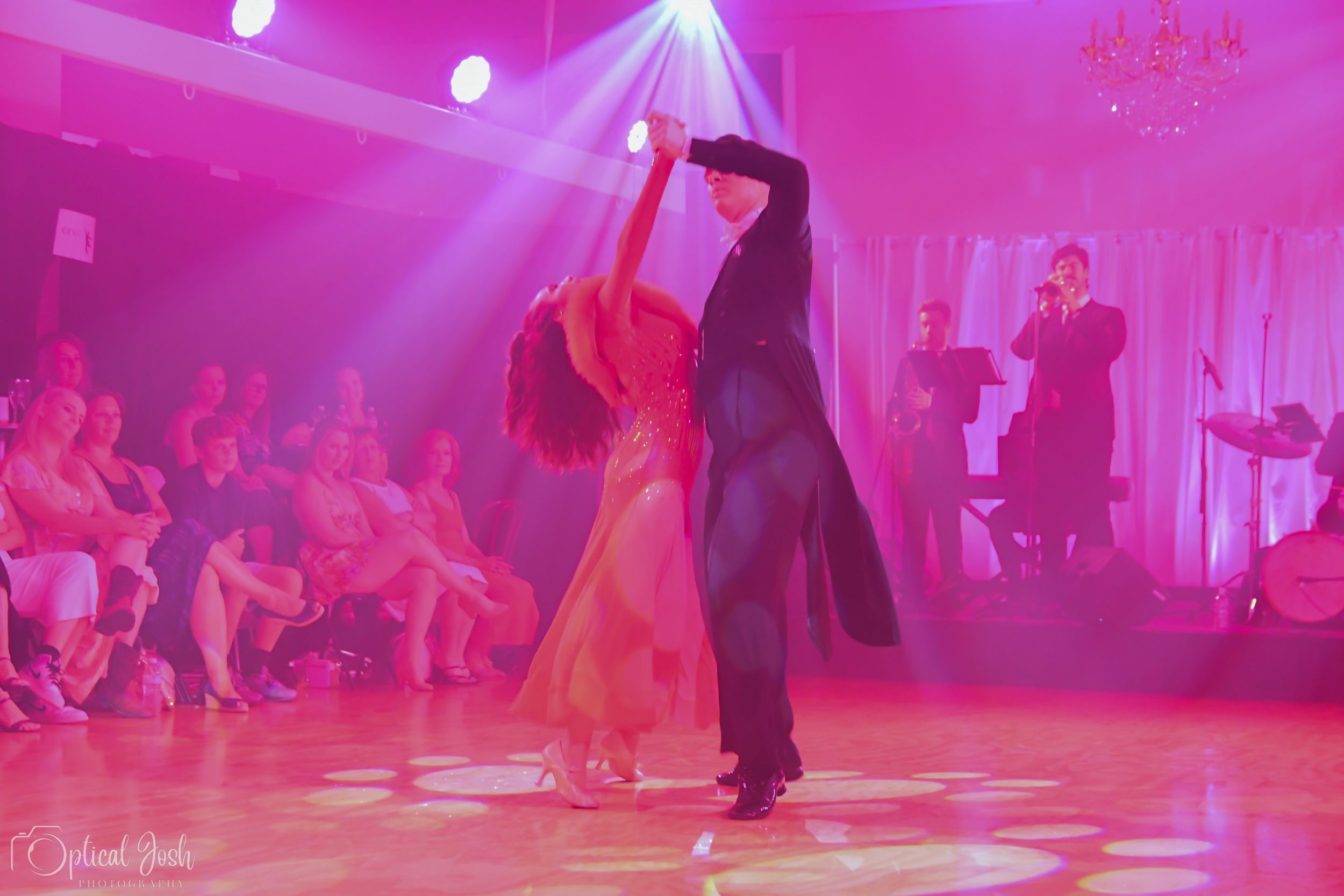 Dance partners perform elegant dance moves under magenta stage lighting with audience in background.