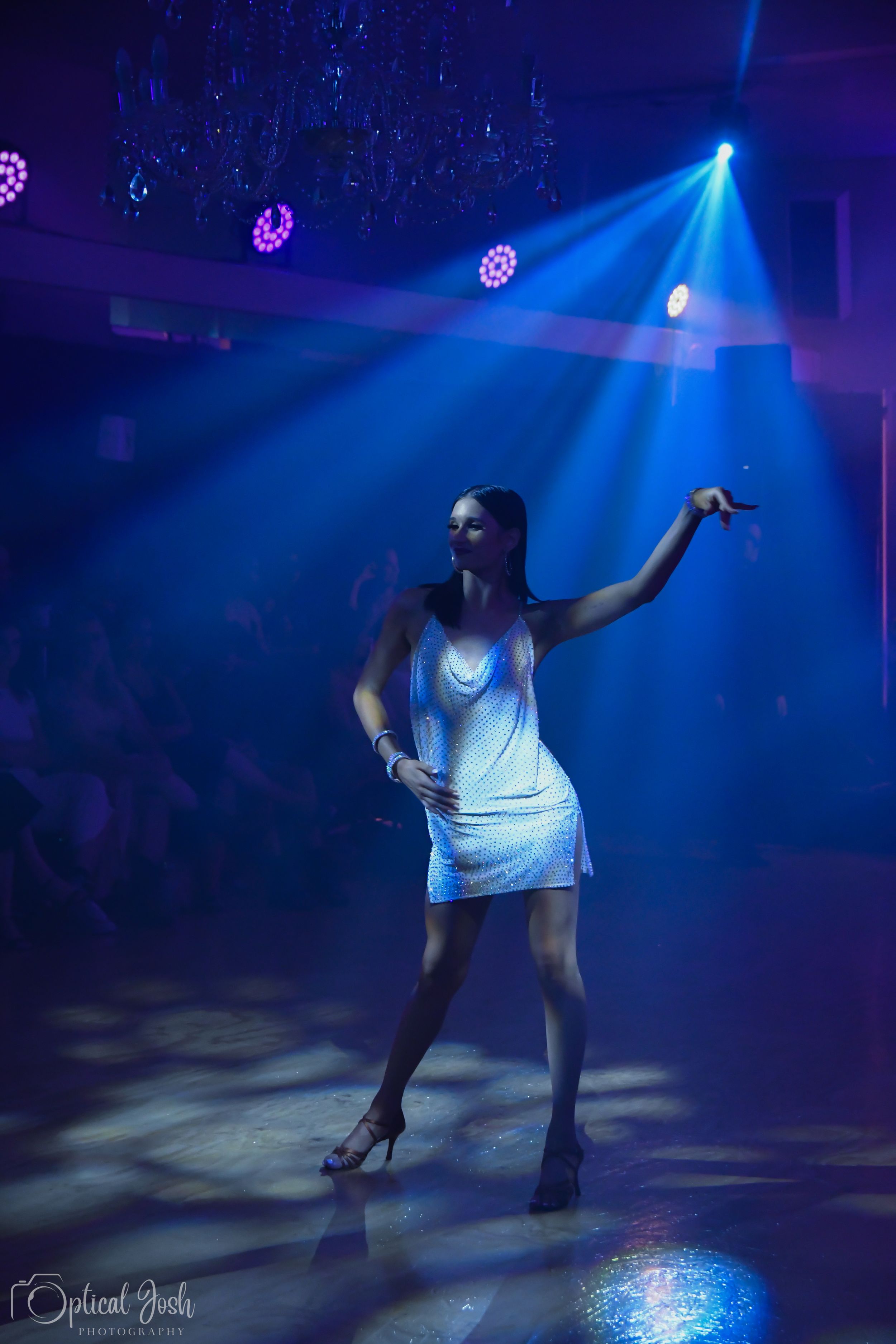 Performer in a white dress dancing under dramatic blue stage lighting and disco balls at a nightclub.