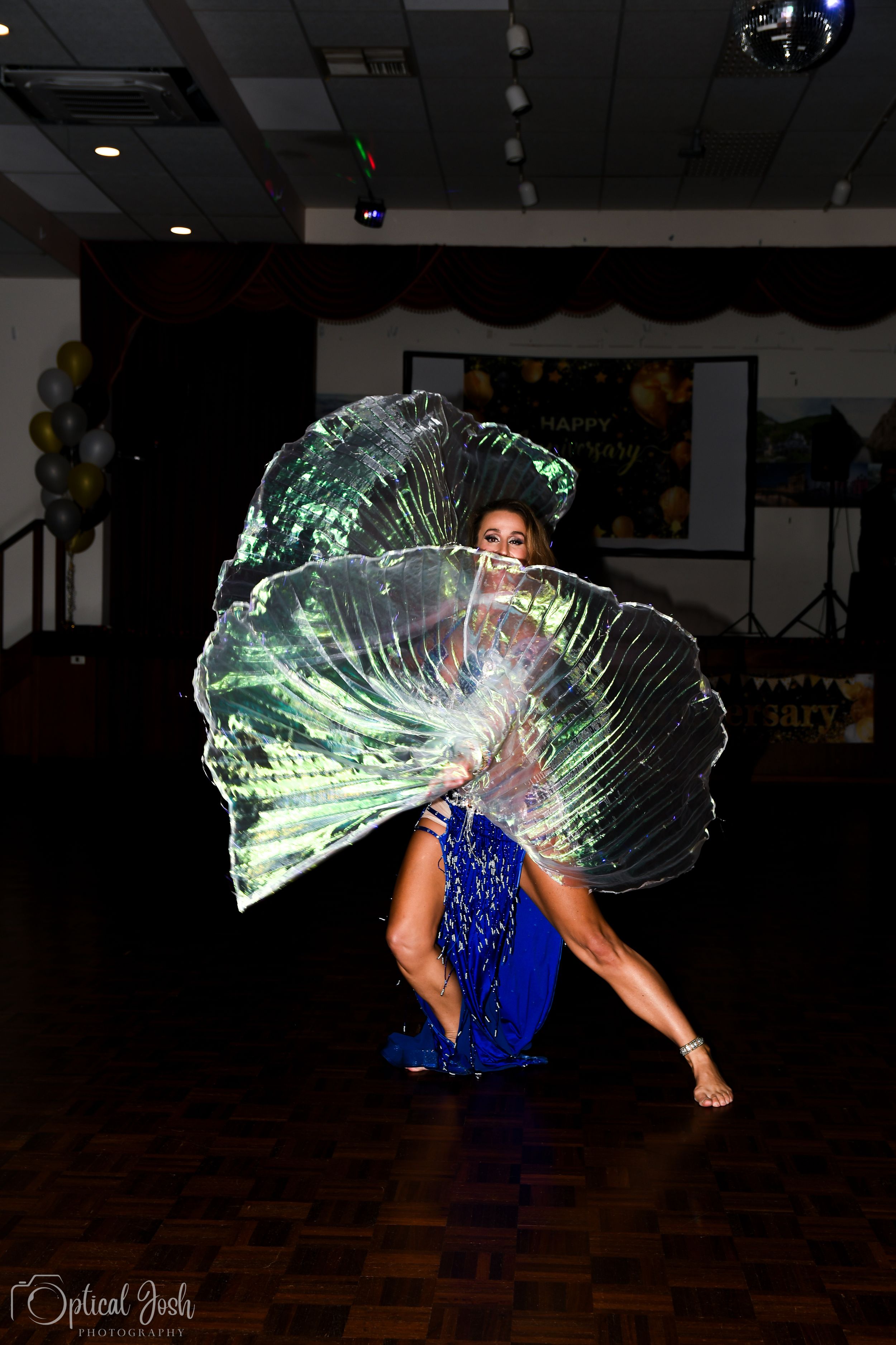 Performer in a sparkling blue dress creates dramatic effect with illuminated translucent fan-like props on dark dance floor.