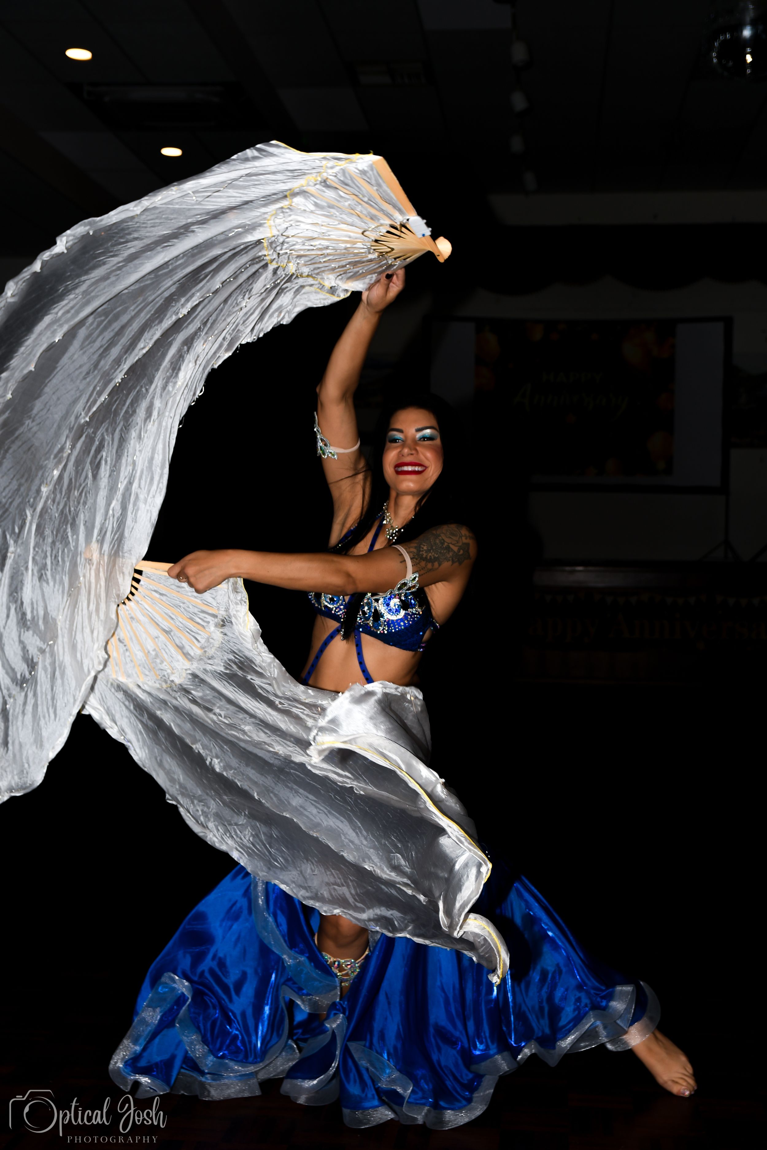 A belly dancer in a sparkly blue costume performs with flowing white silk veils against a dark background.