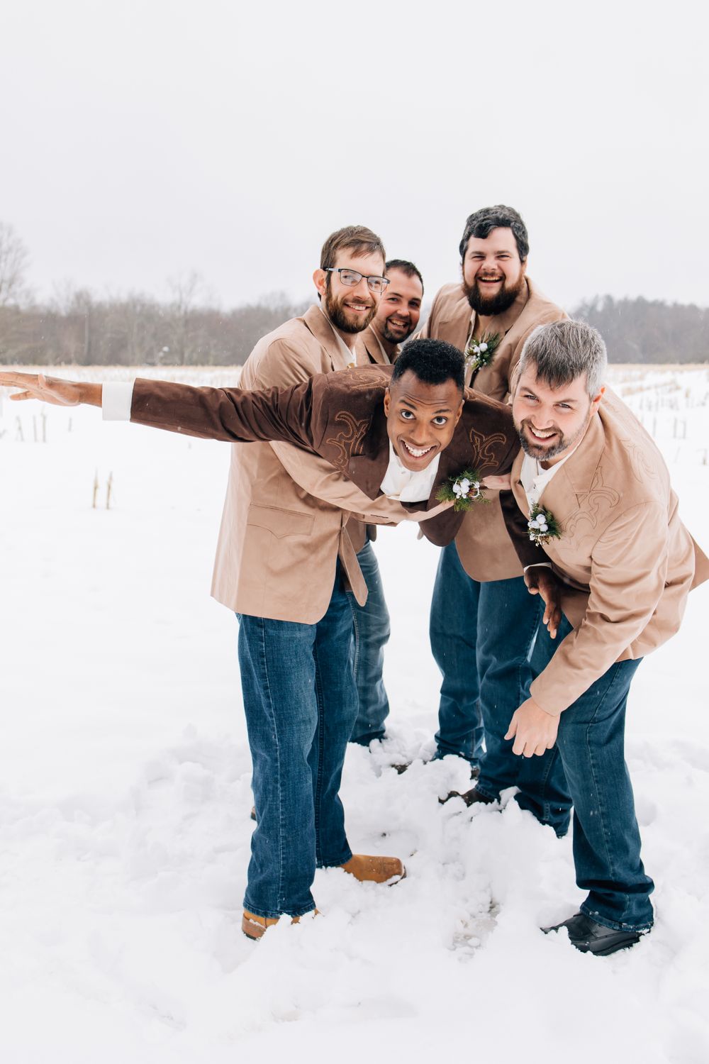 Groomsmen in tan jackets playfully posing together in the snow.