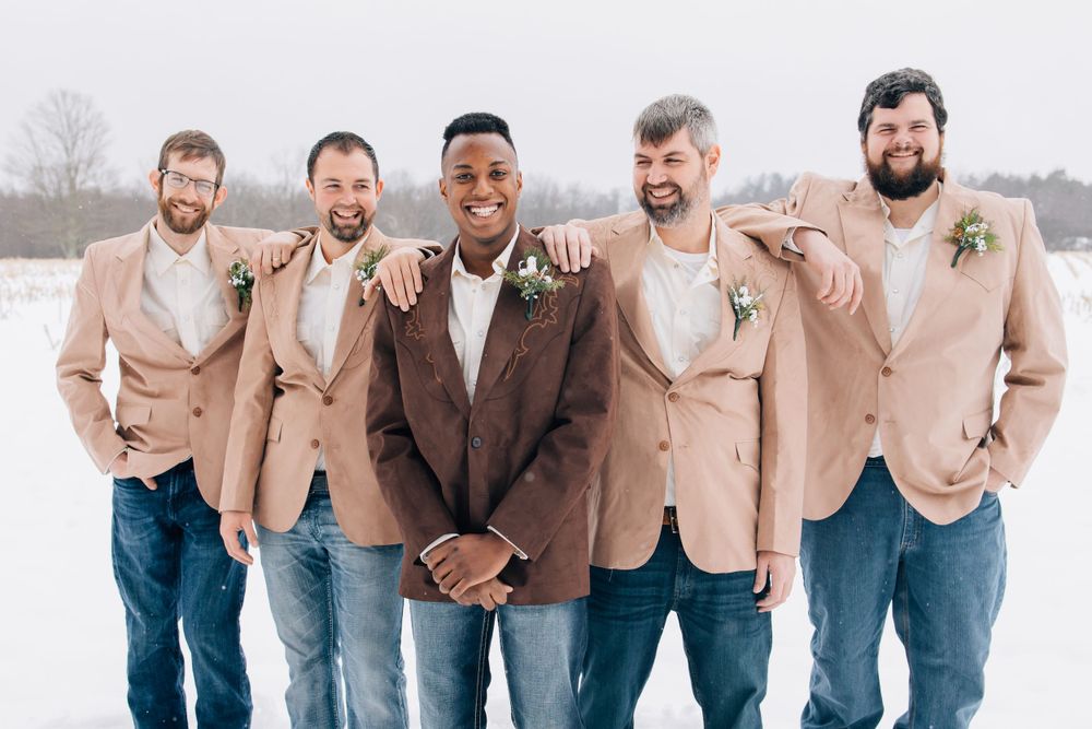 Wedding party groomsmen wearing beige blazers and jeans pose together in winter setting.