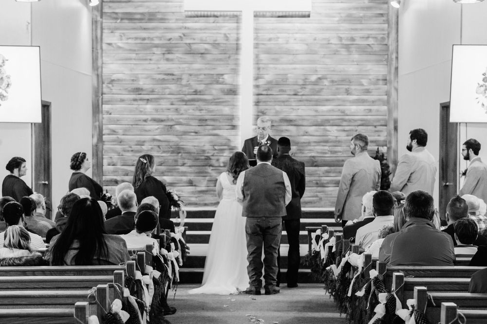 Wedding ceremony in progress inside rustic chapel with wooden walls and pews filled with guests.