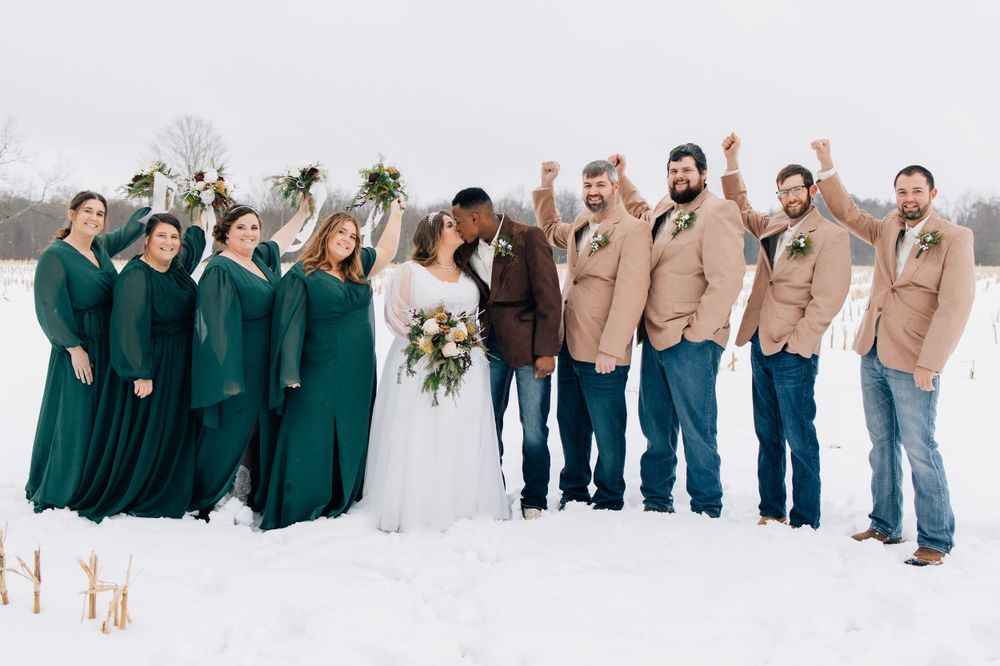 Wedding party in emerald green dresses and tan blazers poses together in snowy landscape.