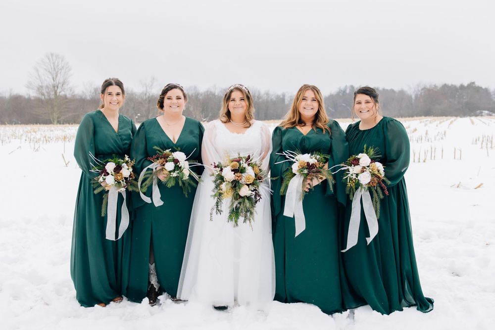 Bridesmaids in emerald green dresses pose with bride in white against snowy landscape.