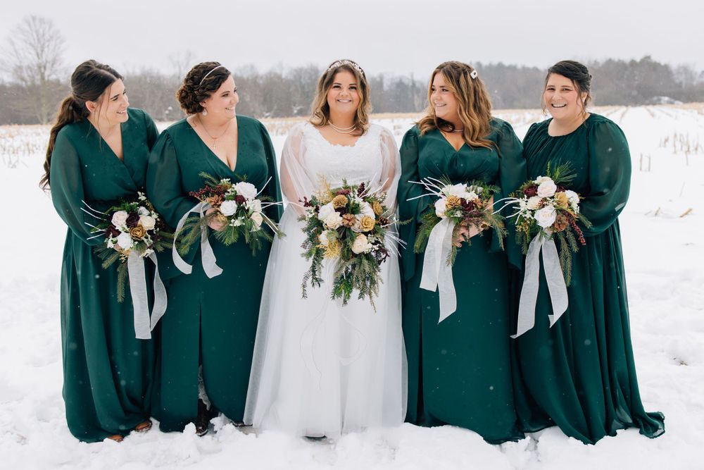 Bridesmaids in emerald green dresses stand with bride holding winter bouquets in snow.