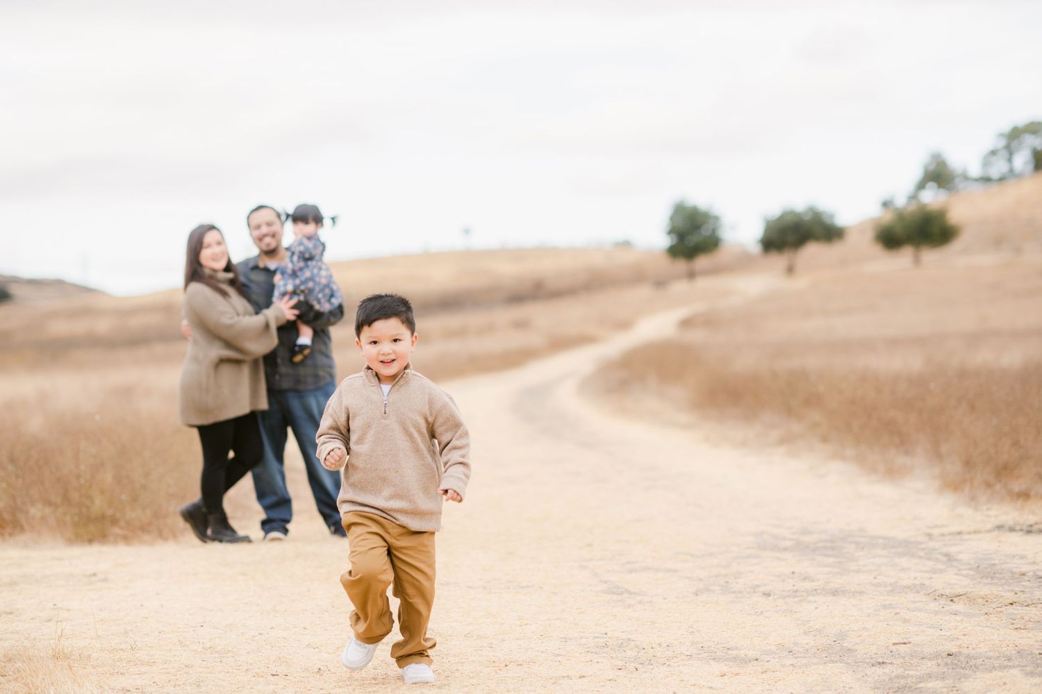 A family walks together on a dirt path through golden rolling hills on a sunny day.
